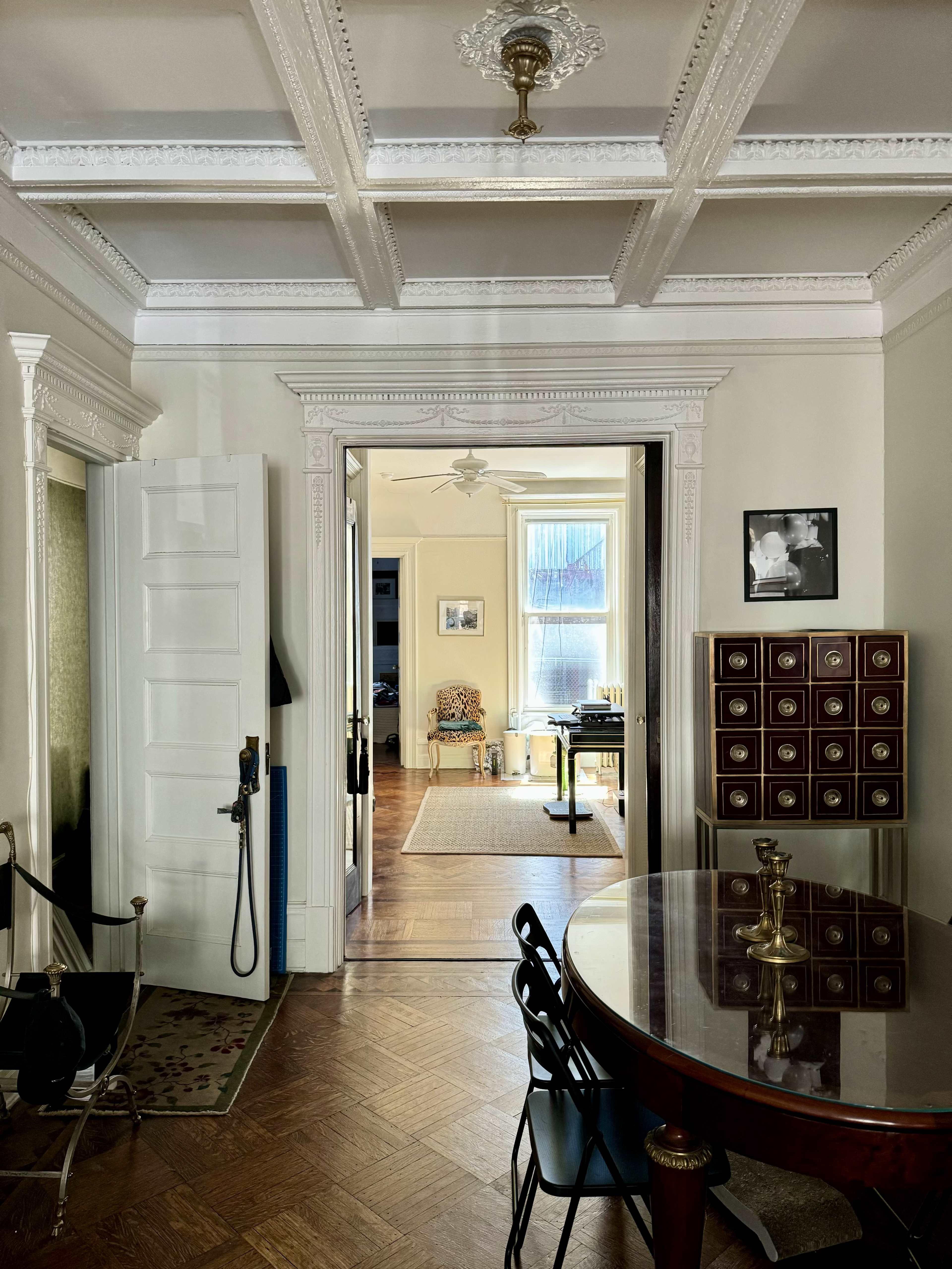 A well-lit hallway with a doorway leading into a spacious room, framed by ornate ceiling details and furniture arranged in a classic style.