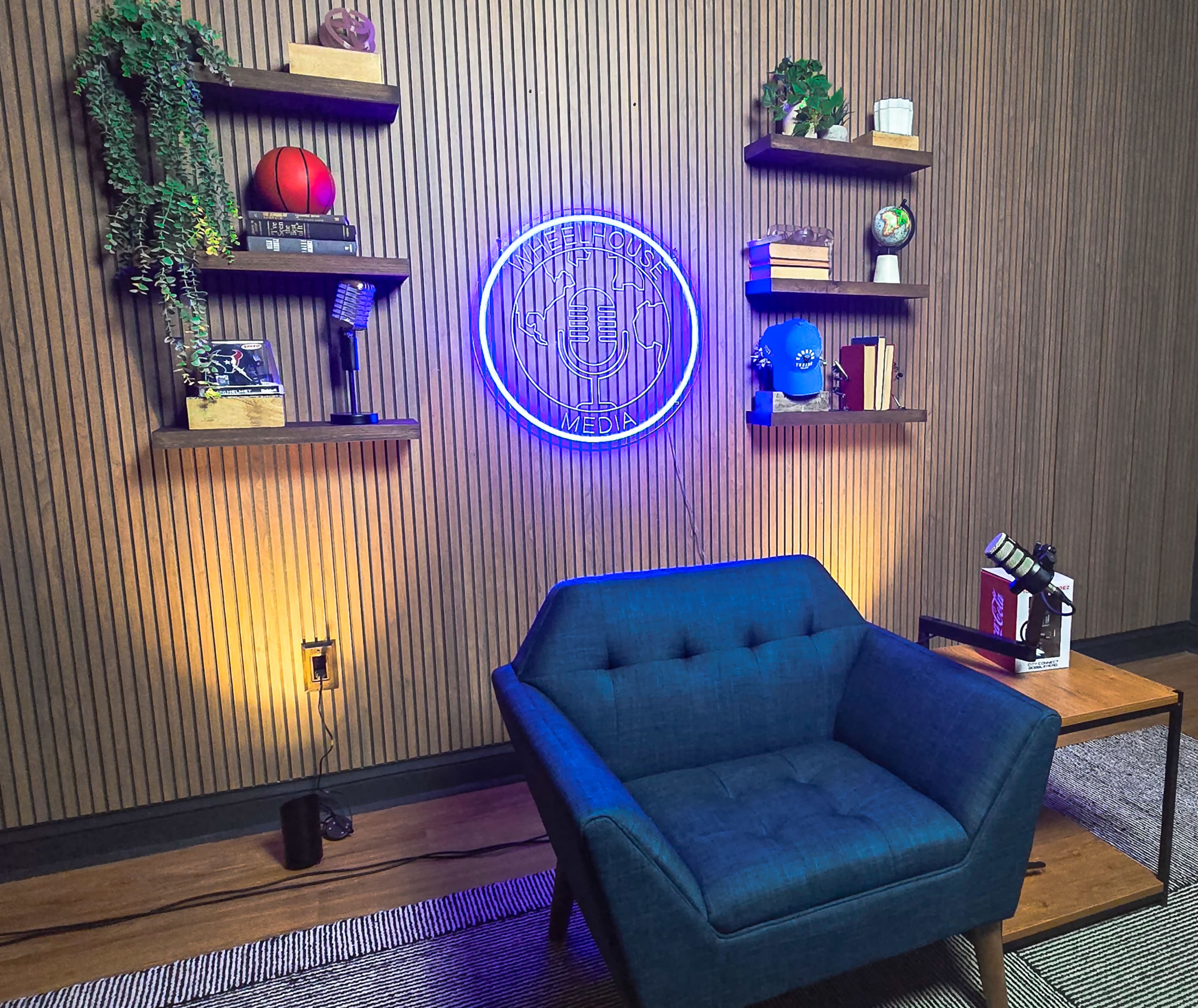 The image shows a cozy media studio setup with a blue armchair facing a wall adorned with shelves, a neon sign, and various decorative items.