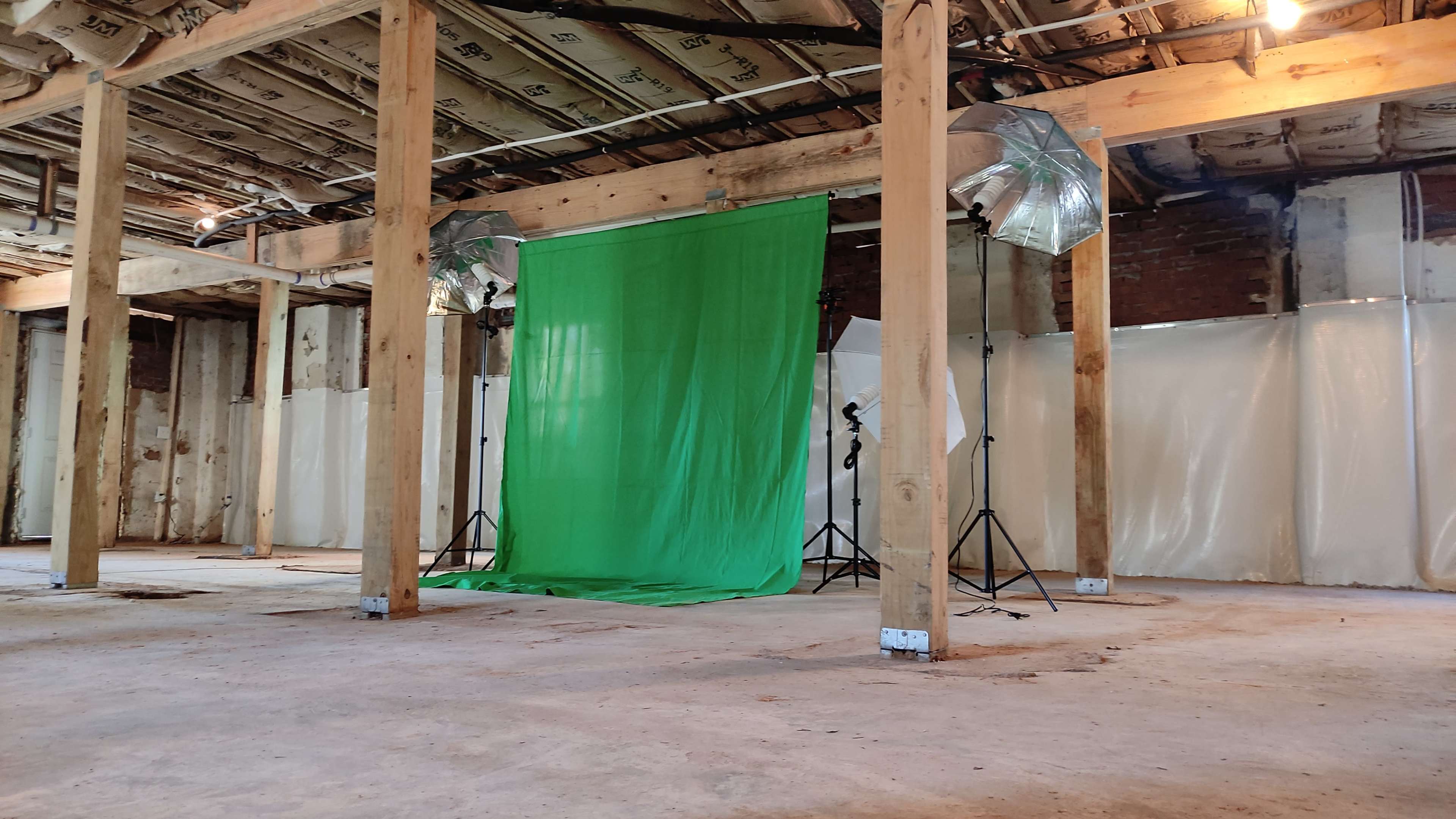 An empty room with wooden beams and a green backdrop set up for photography, flanked by lighting equipment on tripods.