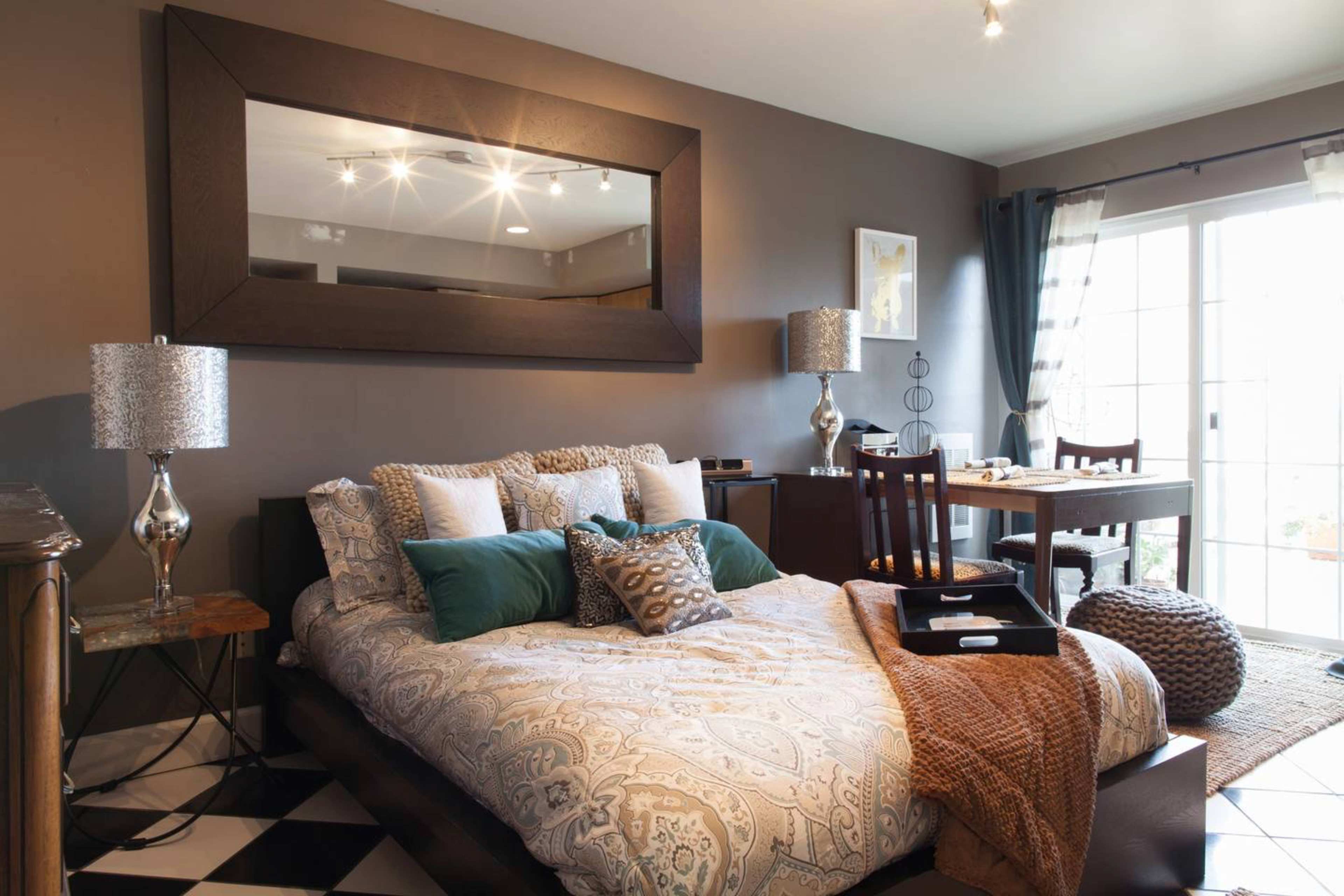A modern bedroom featuring a bed with patterned bedding, two bedside lamps, a large mirror, and a small dining area with a wooden table and chairs.