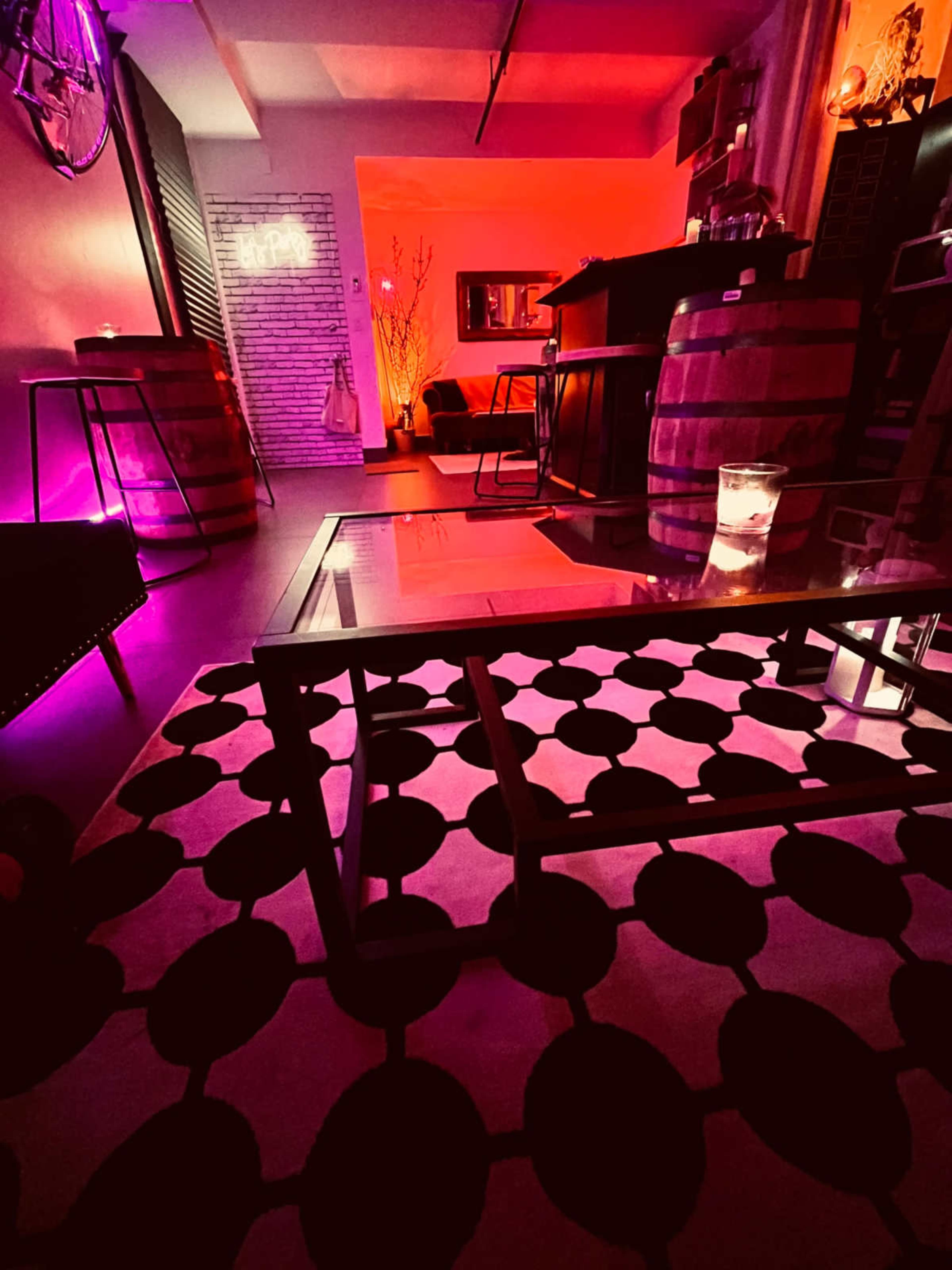 The image shows a stylish interior space with a patterned black and white rug, a glass coffee table, and wooden barrels serving as decor, illuminated by red and purple lighting.