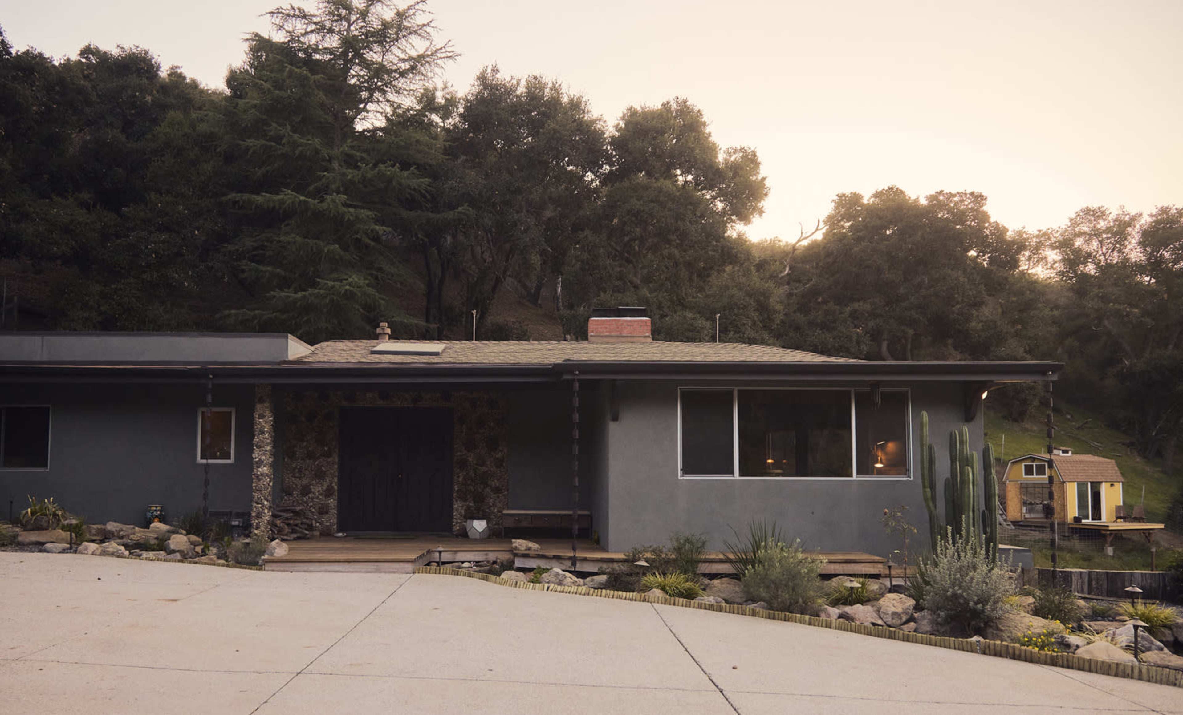 A modern house with a stone accent wall is set on a sloped landscape, surrounded by greenery and accompanied by a small shed in the background.