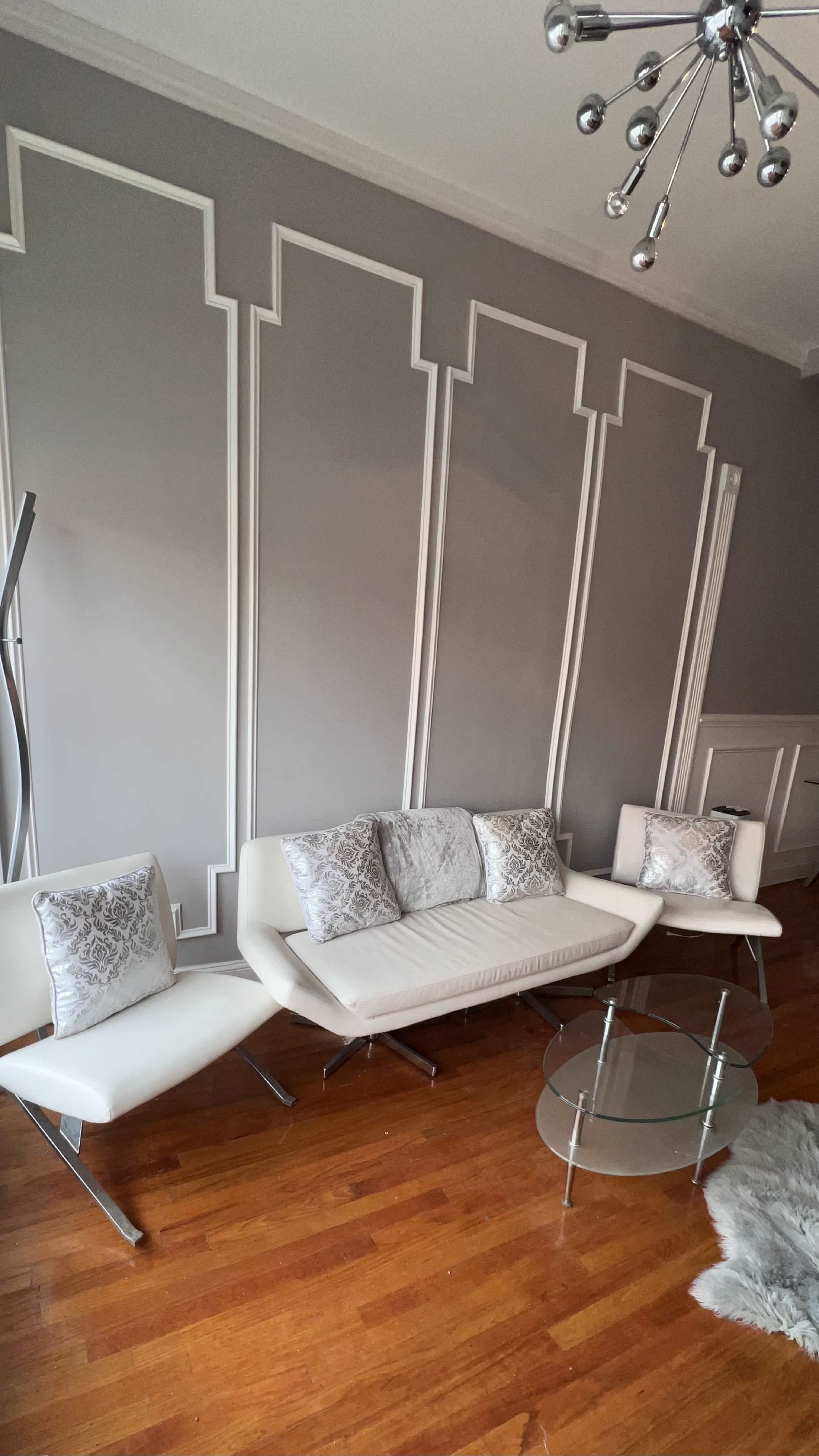 The image shows a modern living room featuring a white leather sofa and chairs, a glass coffee table, and decorative wall paneling.