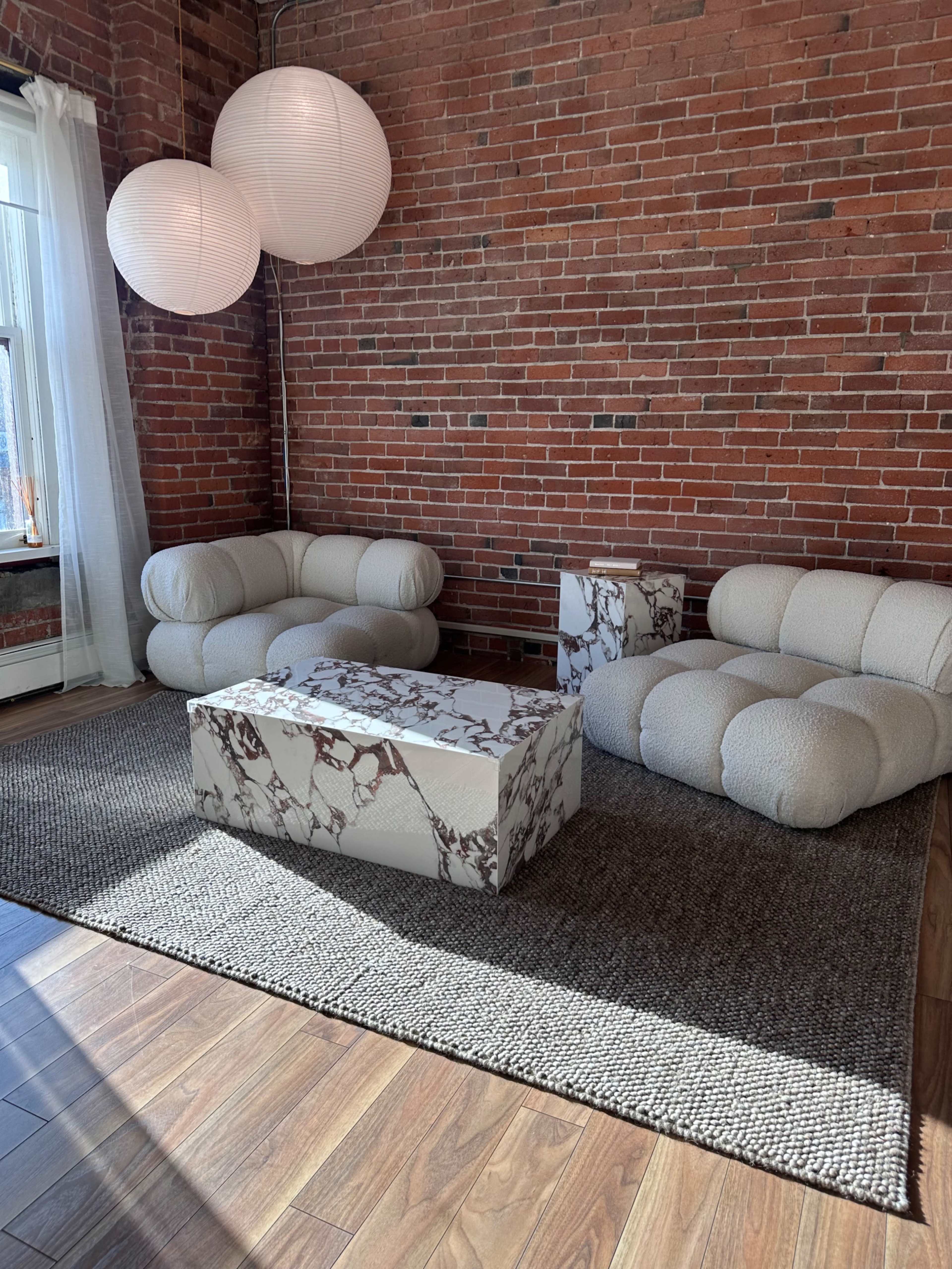 The image shows a cozy living area with two upholstered sofas, a marble-patterned coffee table, and paper lanterns suspended from the ceiling, all set against a brick wall.