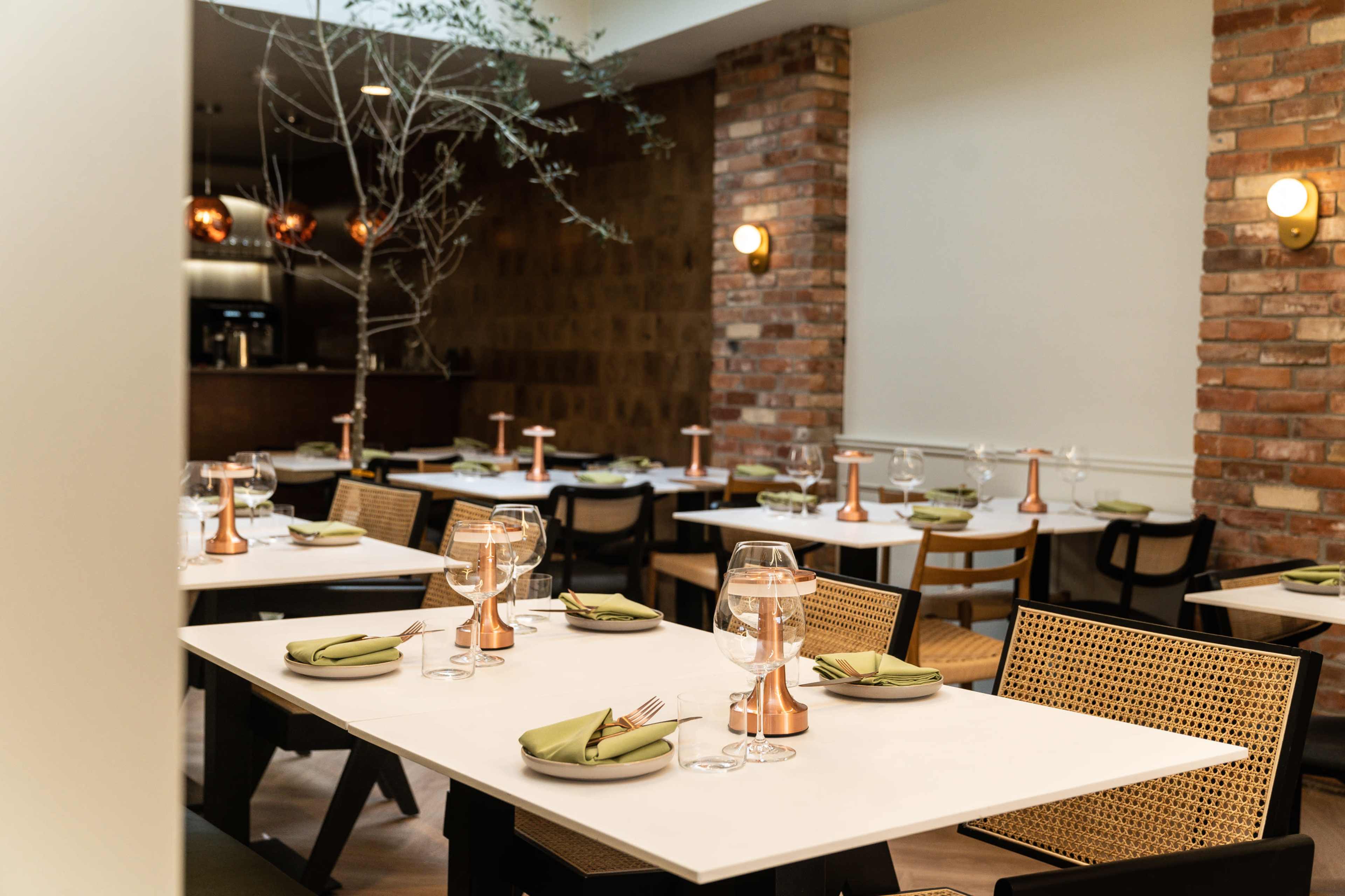 A modern dining space features tables set with green napkins, glassware, and copper accents, complemented by brick walls and soft lighting.