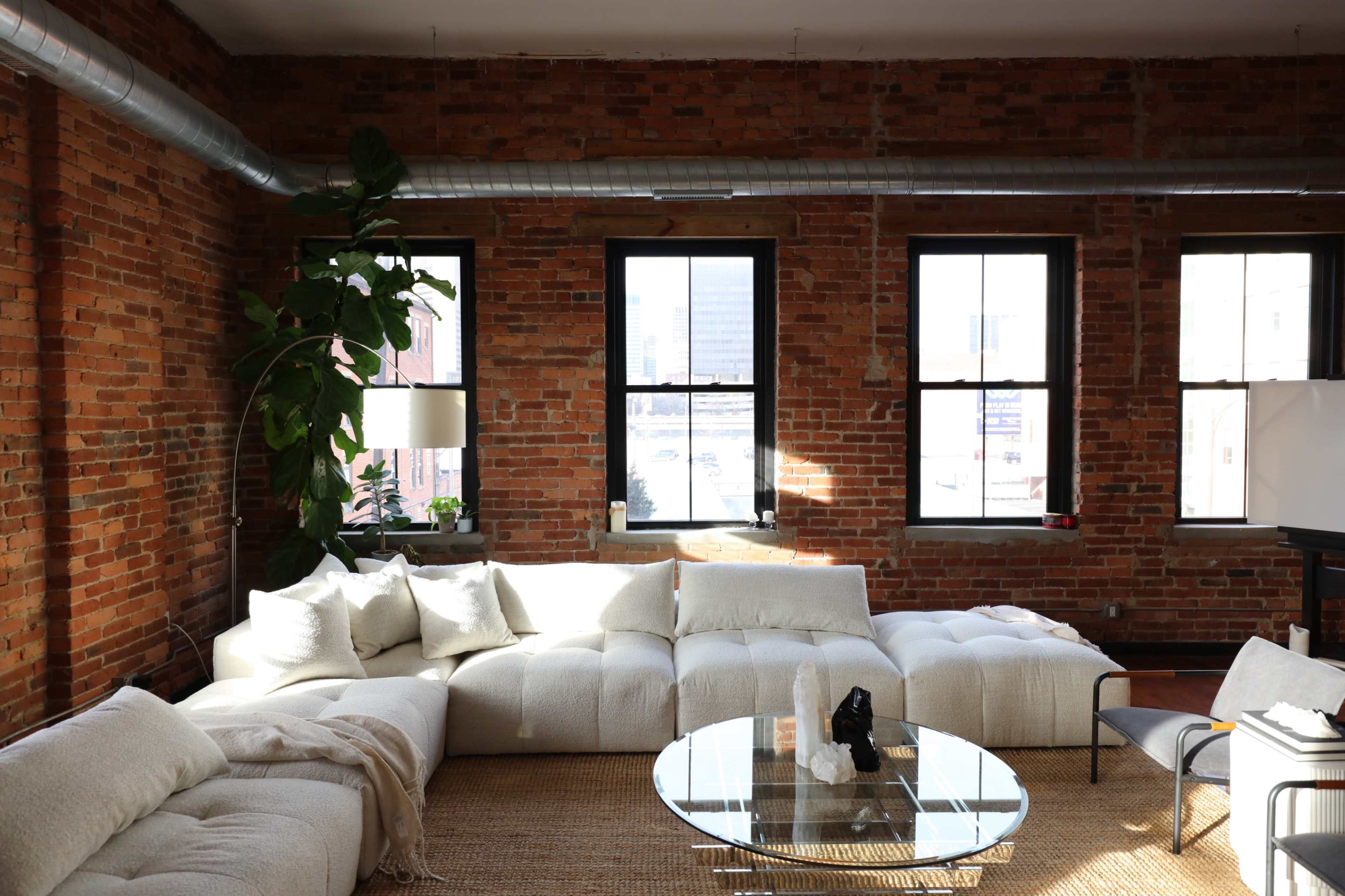 A spacious living room features a large, white sectional sofa, a glass coffee table, and several windows that provide natural light against exposed brick walls.