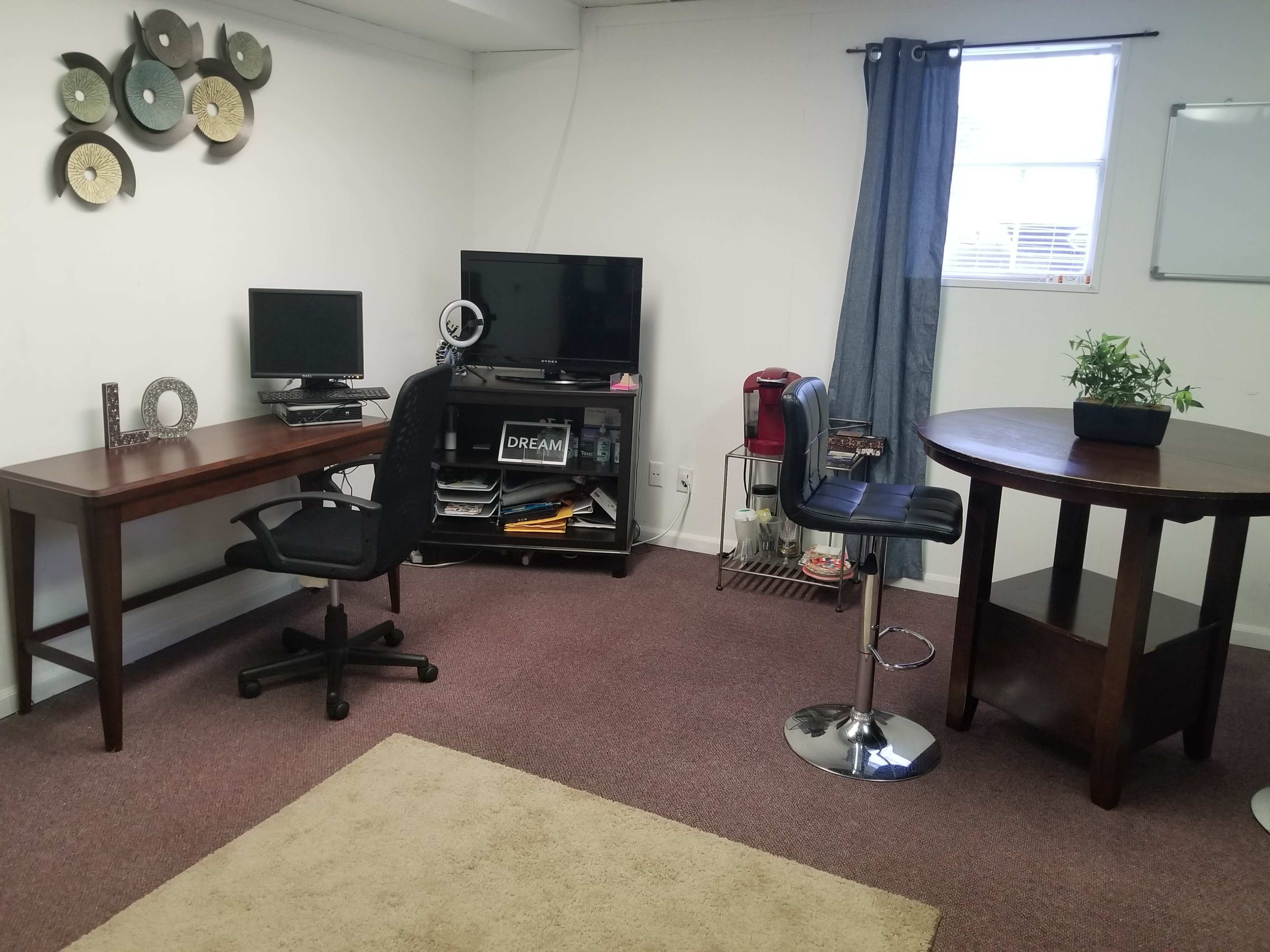 A small office space with two desks, a computer setup, a television, a coffee maker, and a round table with a plant on it.