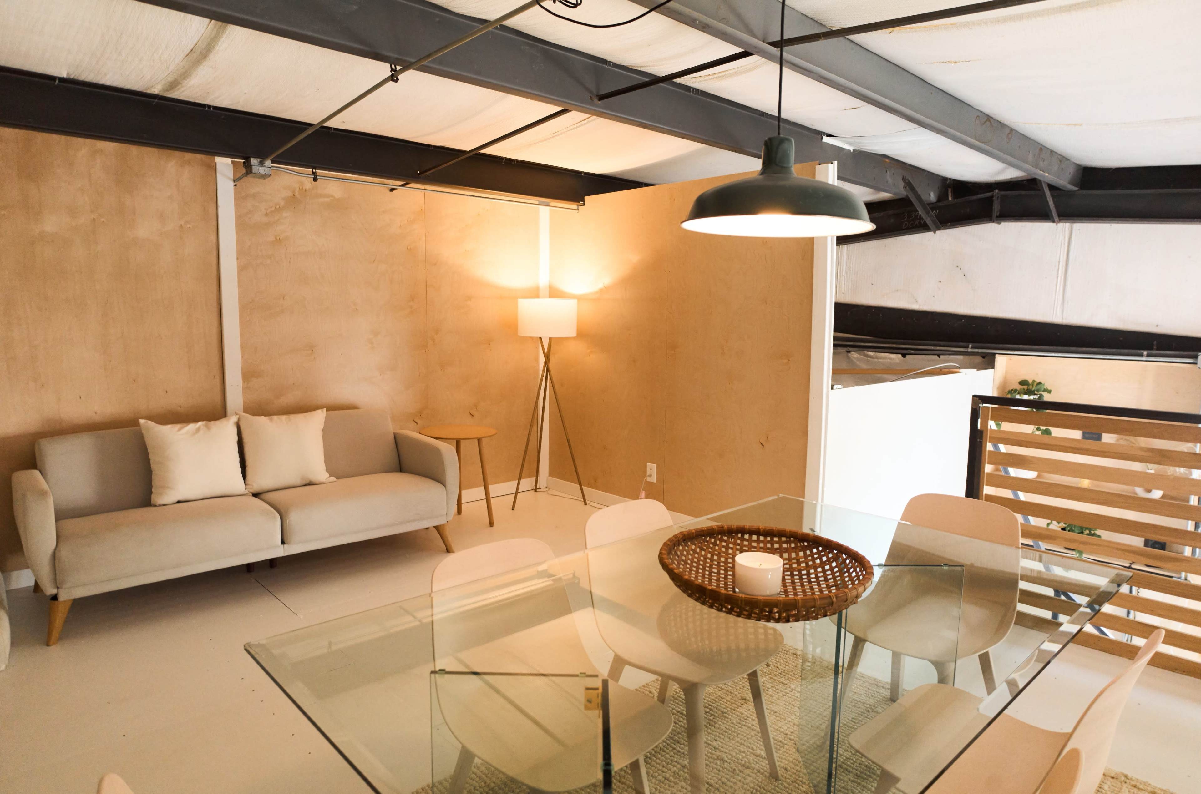 The image features a modern living space with a light-colored sofa, a lamp, a glass dining table surrounded by white chairs, and wooden paneling on the walls.