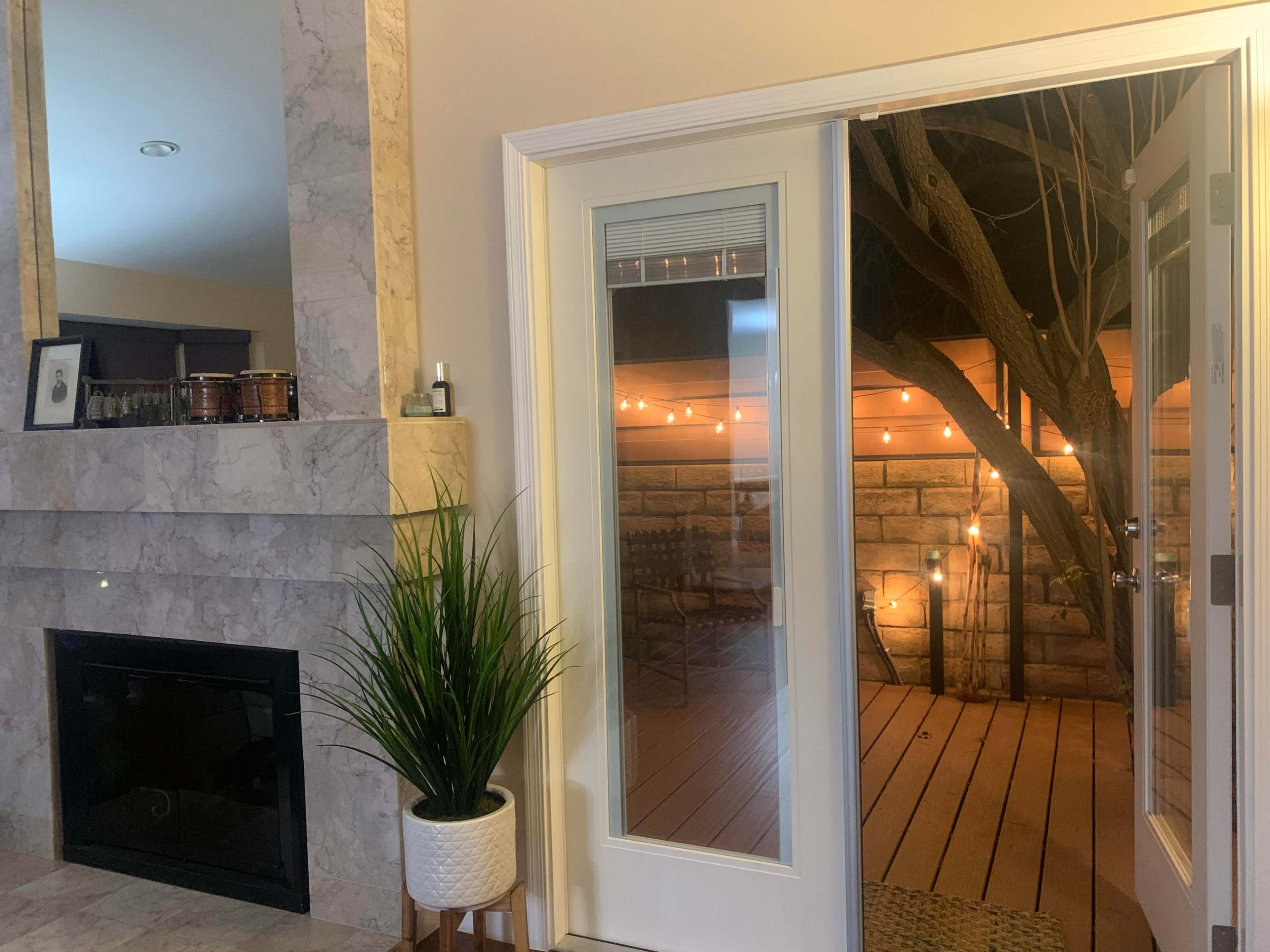 The image shows an interior living space with a fireplace on the left and a set of glass doors leading to a wooden deck illuminated by string lights.