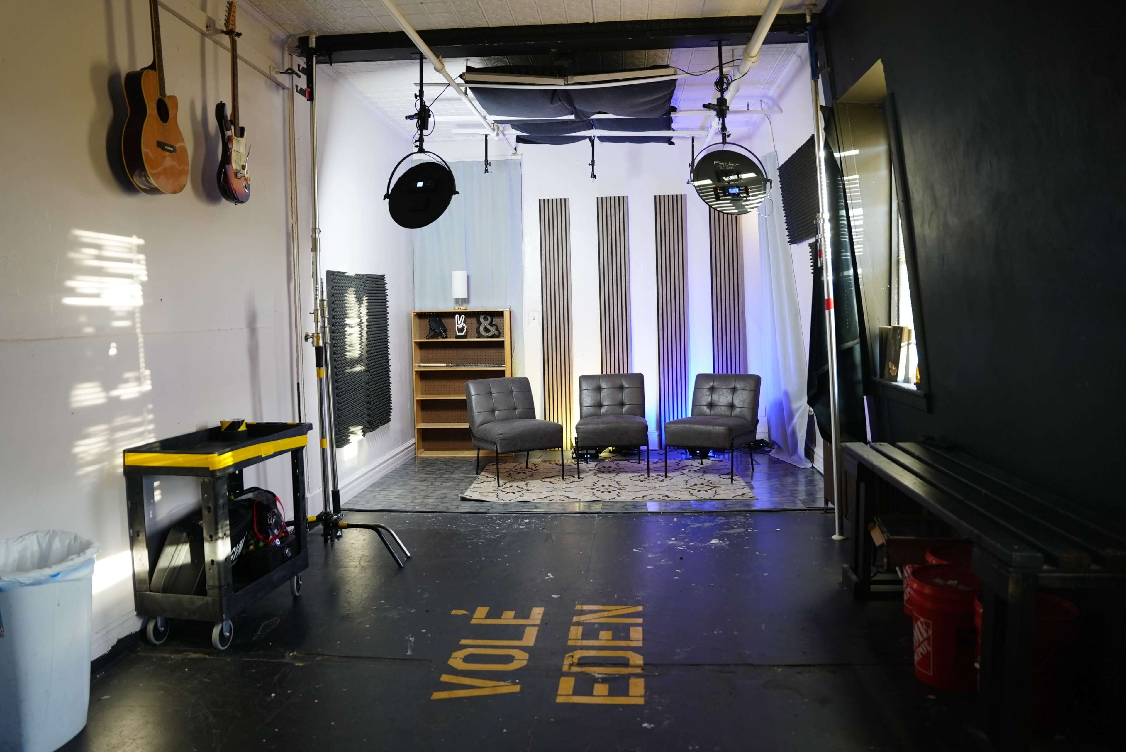 The image shows a studio space with acoustic panels, three gray chairs, a bookshelf, and various lighting fixtures.