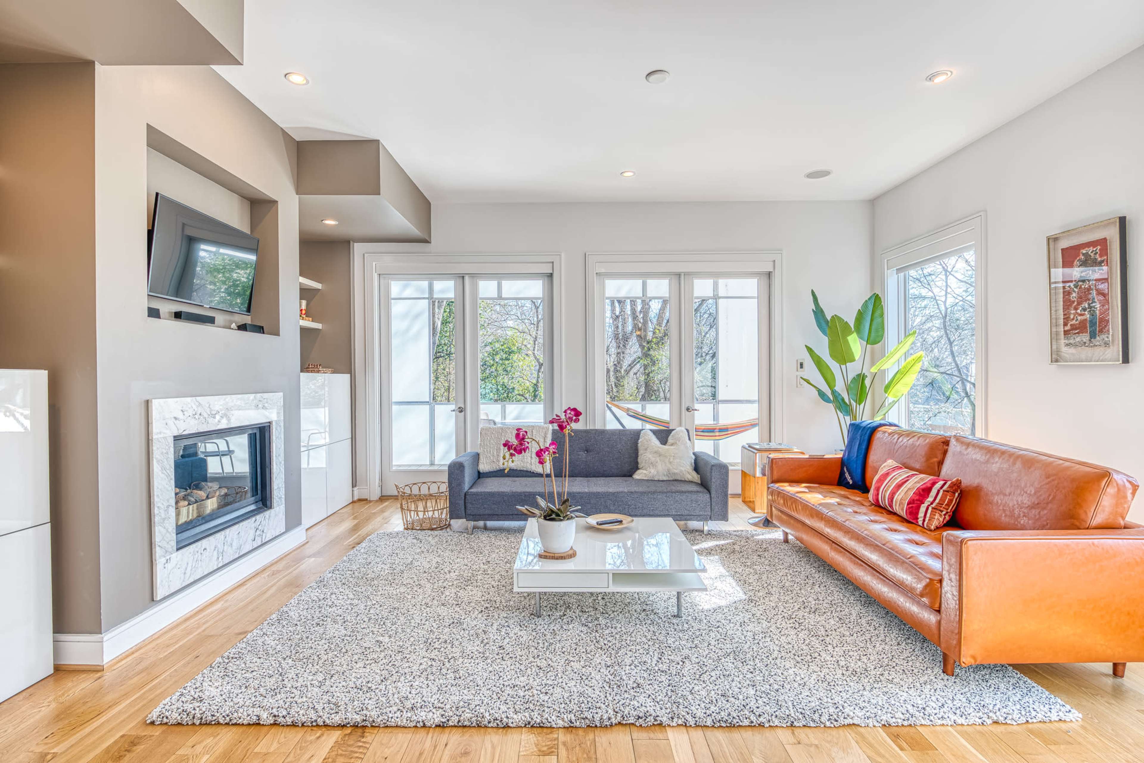 The living room features a gray sofa, an orange leather couch, a coffee table on a textured rug, and large windows allowing natural light.