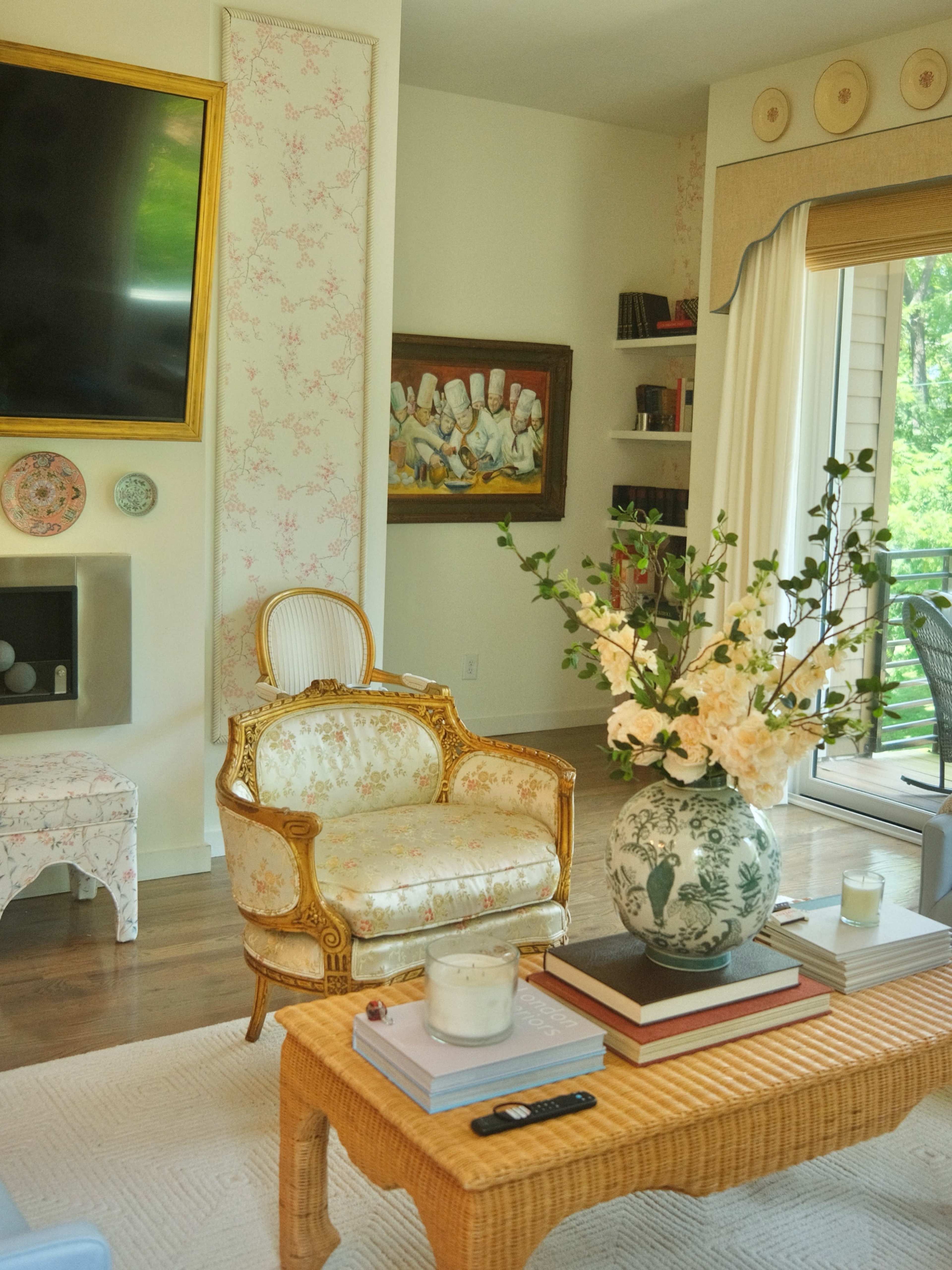 The image shows a neatly decorated living room featuring a floral-patterned chair, a coffee table with books and candles, and large windows with a view of greenery outside.