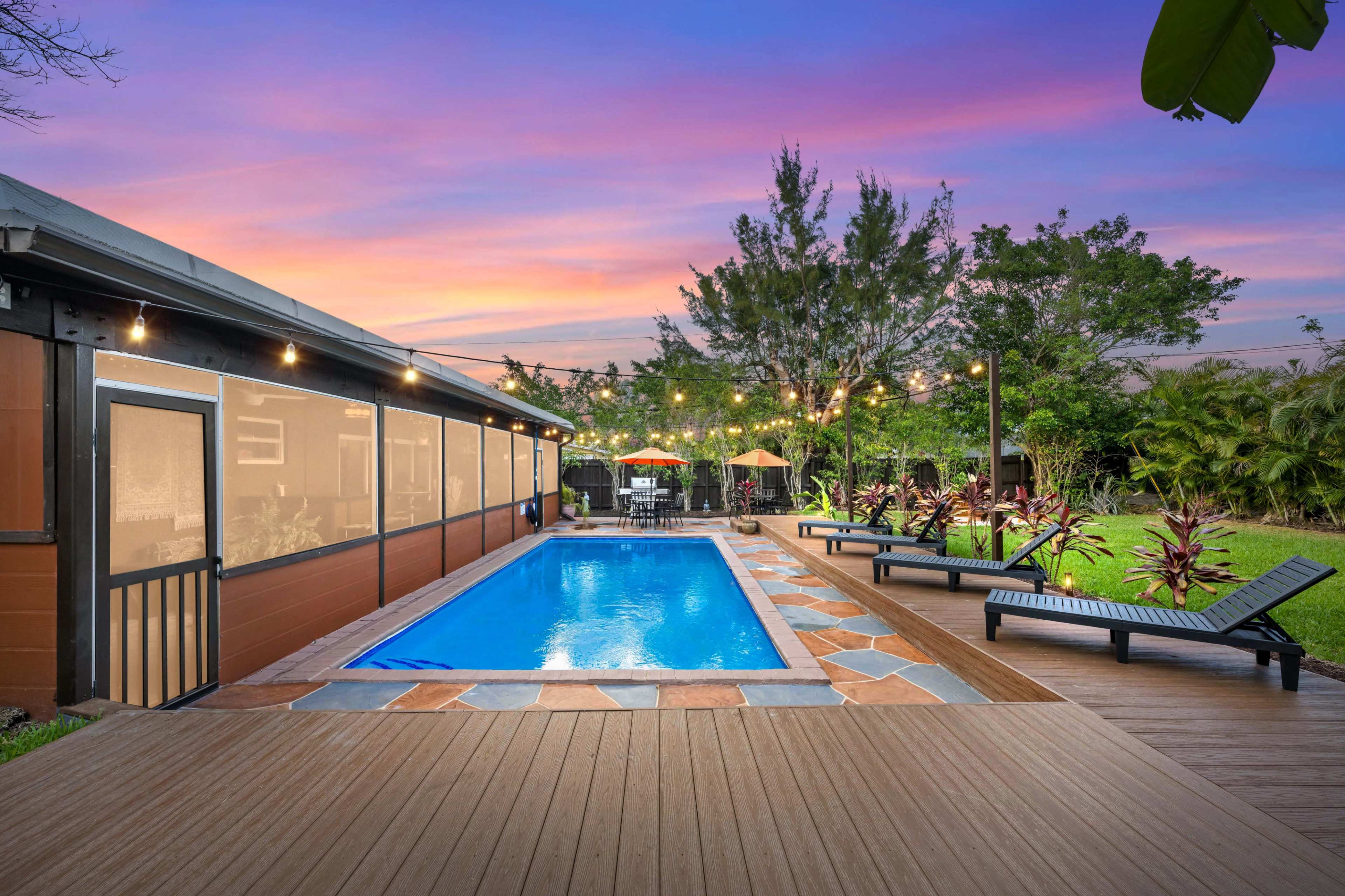 A well-maintained backyard with a rectangular swimming pool surrounded by lounge chairs, a wooden deck, and landscaped foliage, set against a colorful sunset sky.