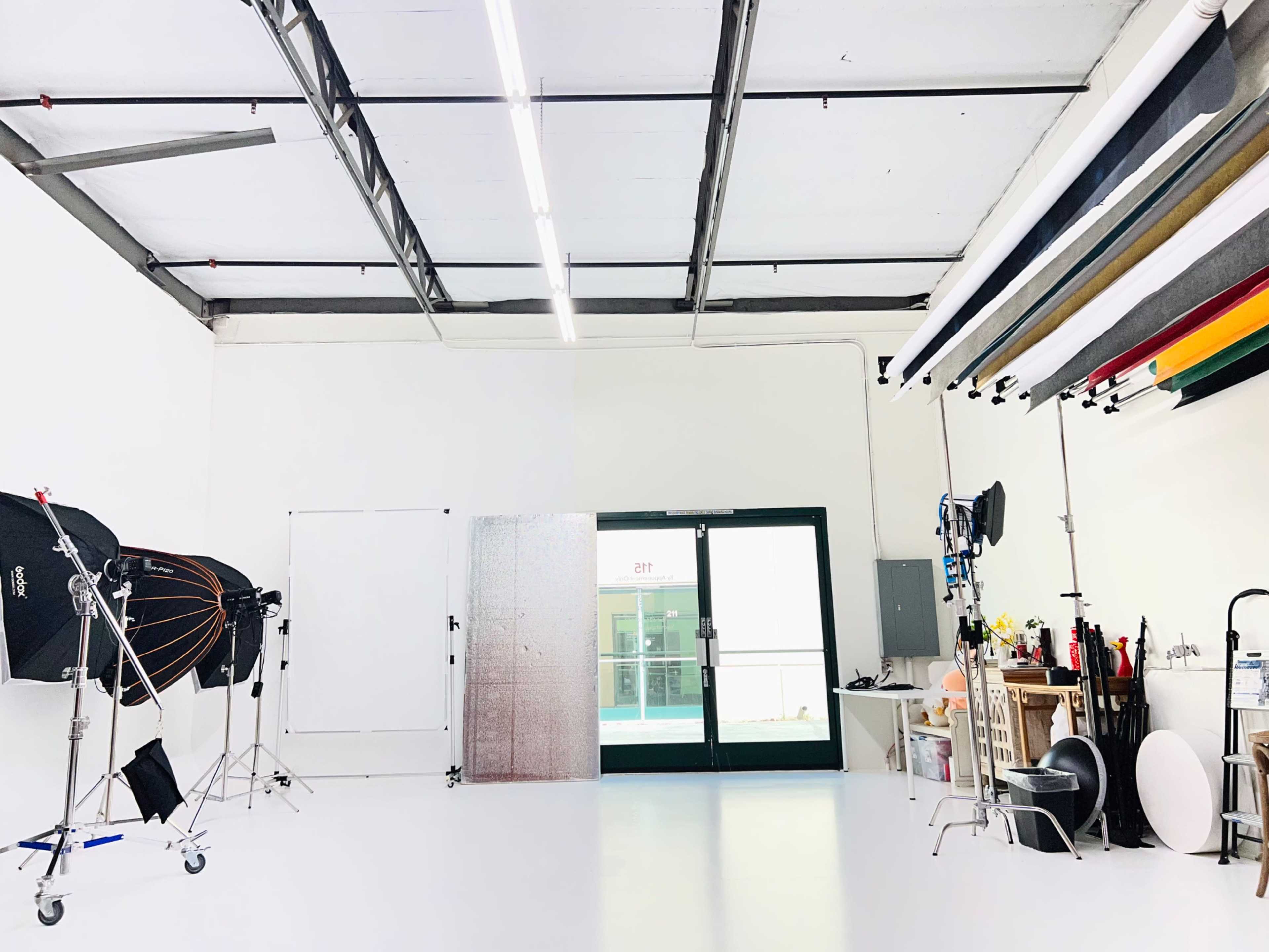 The image shows a photography studio with various lighting equipment, a large open door, and colorful backdrops hanging from the ceiling.