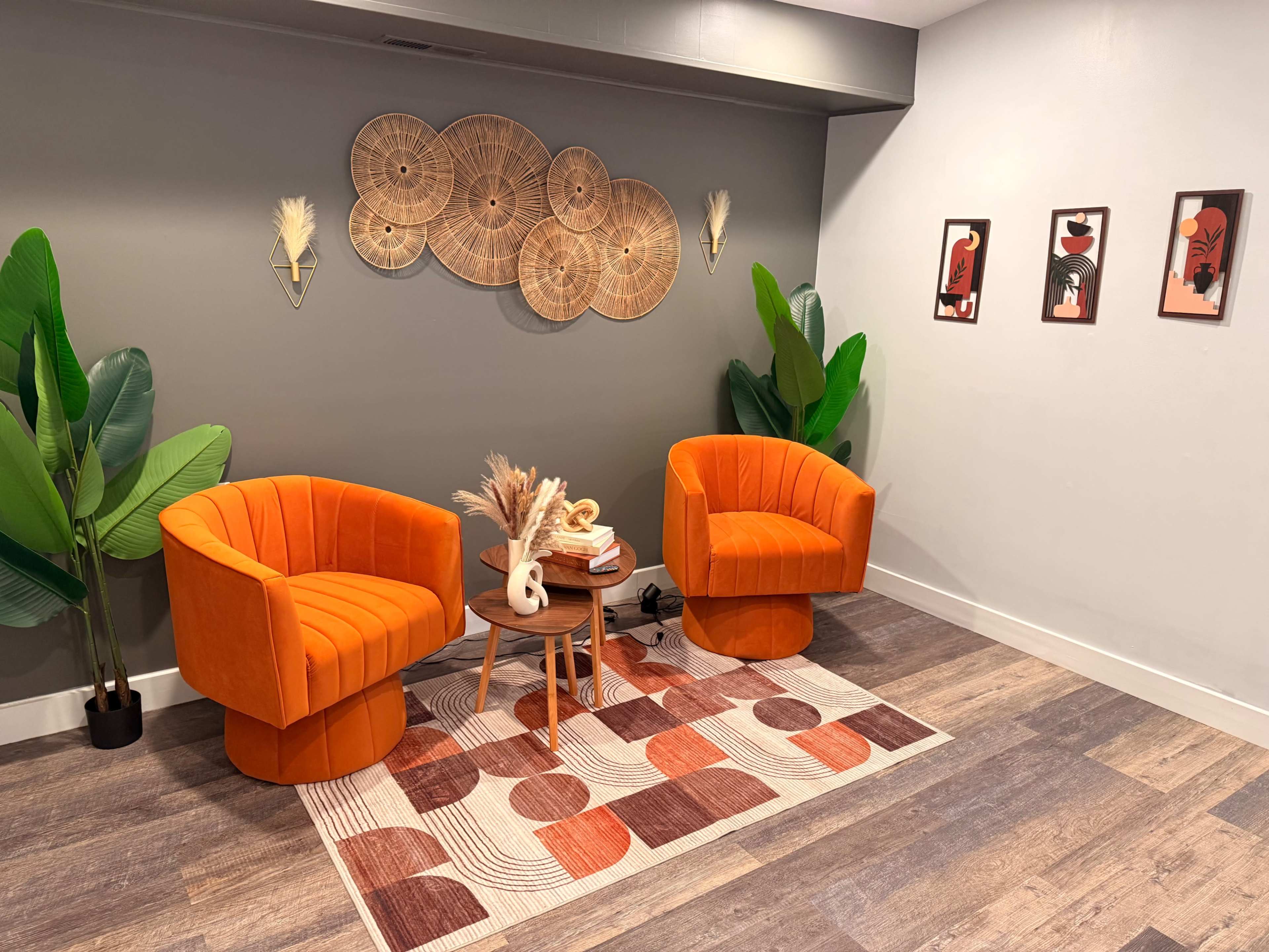 The image shows a cozy interior setting with two orange upholstered chairs, a small round table, a patterned rug, and decorative wall art.