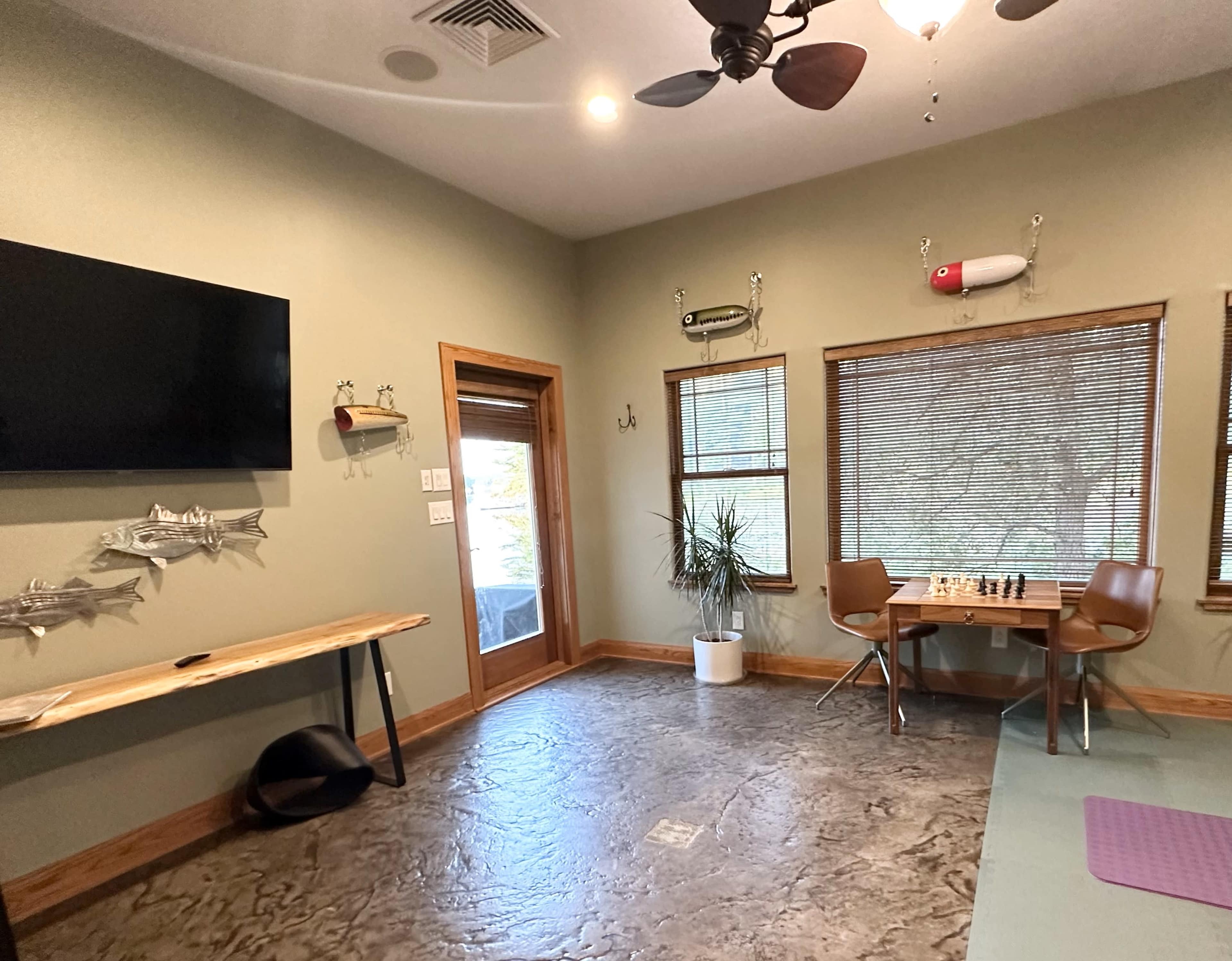The room features a television on the wall, a dining table with chairs, a chess set, and a large window with blinds, all set against a light green wall.