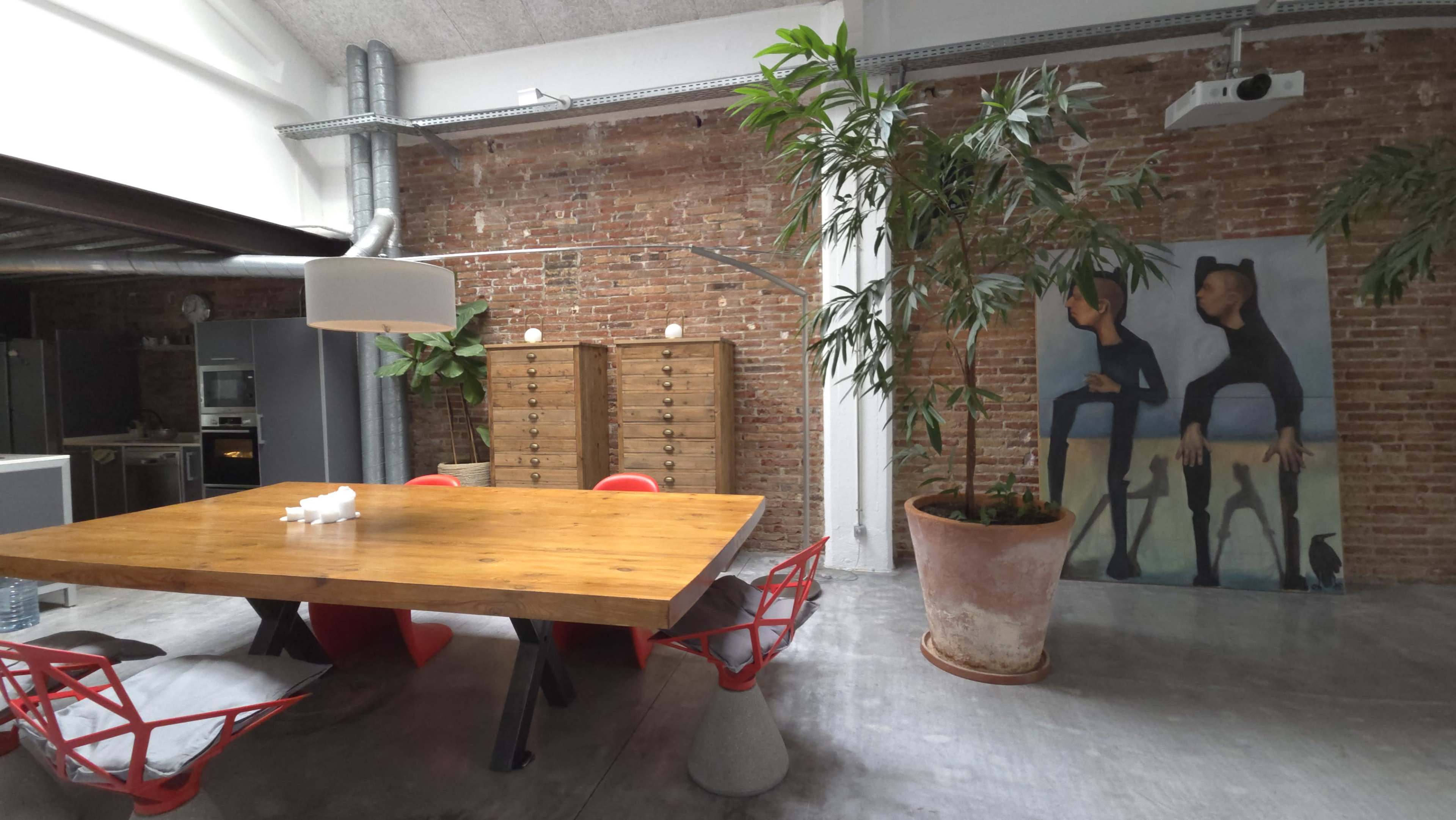 The image shows a modern industrial-style room featuring a wooden dining table surrounded by red chairs, a large potted plant, and a mural on the wall.