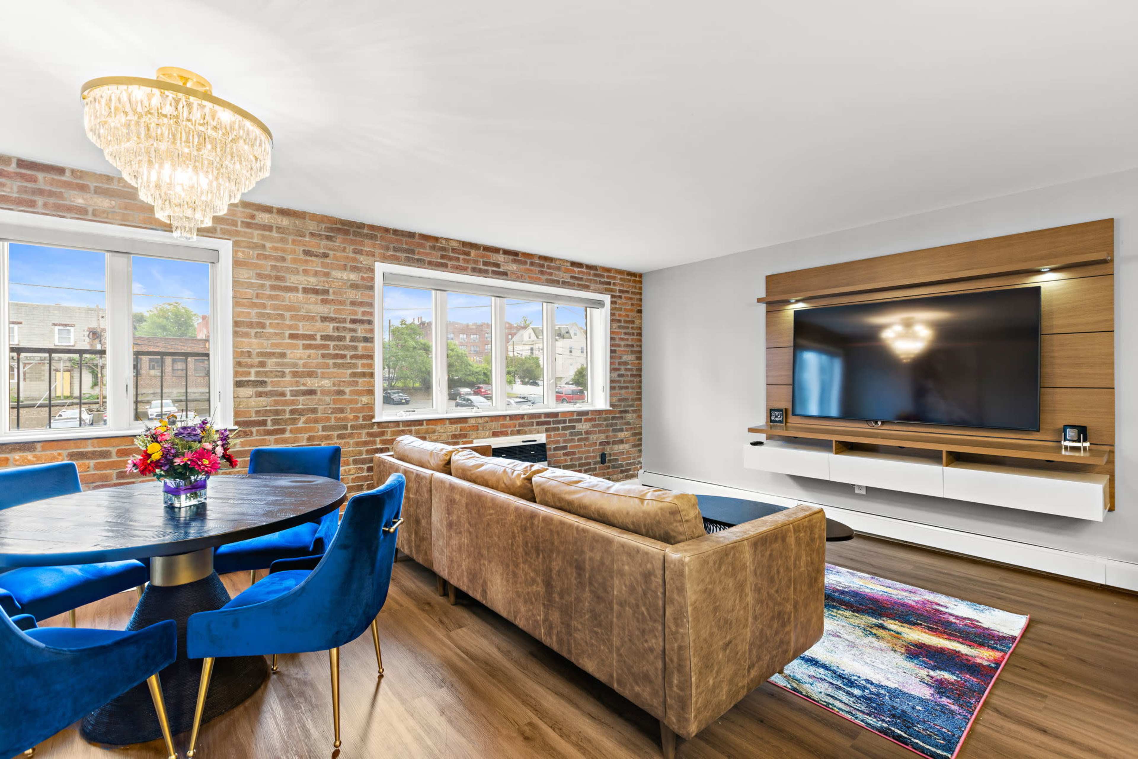 A modern living area with exposed brick walls, a circular dining table with blue chairs, and a large flat-screen TV mounted on a wooden panel.