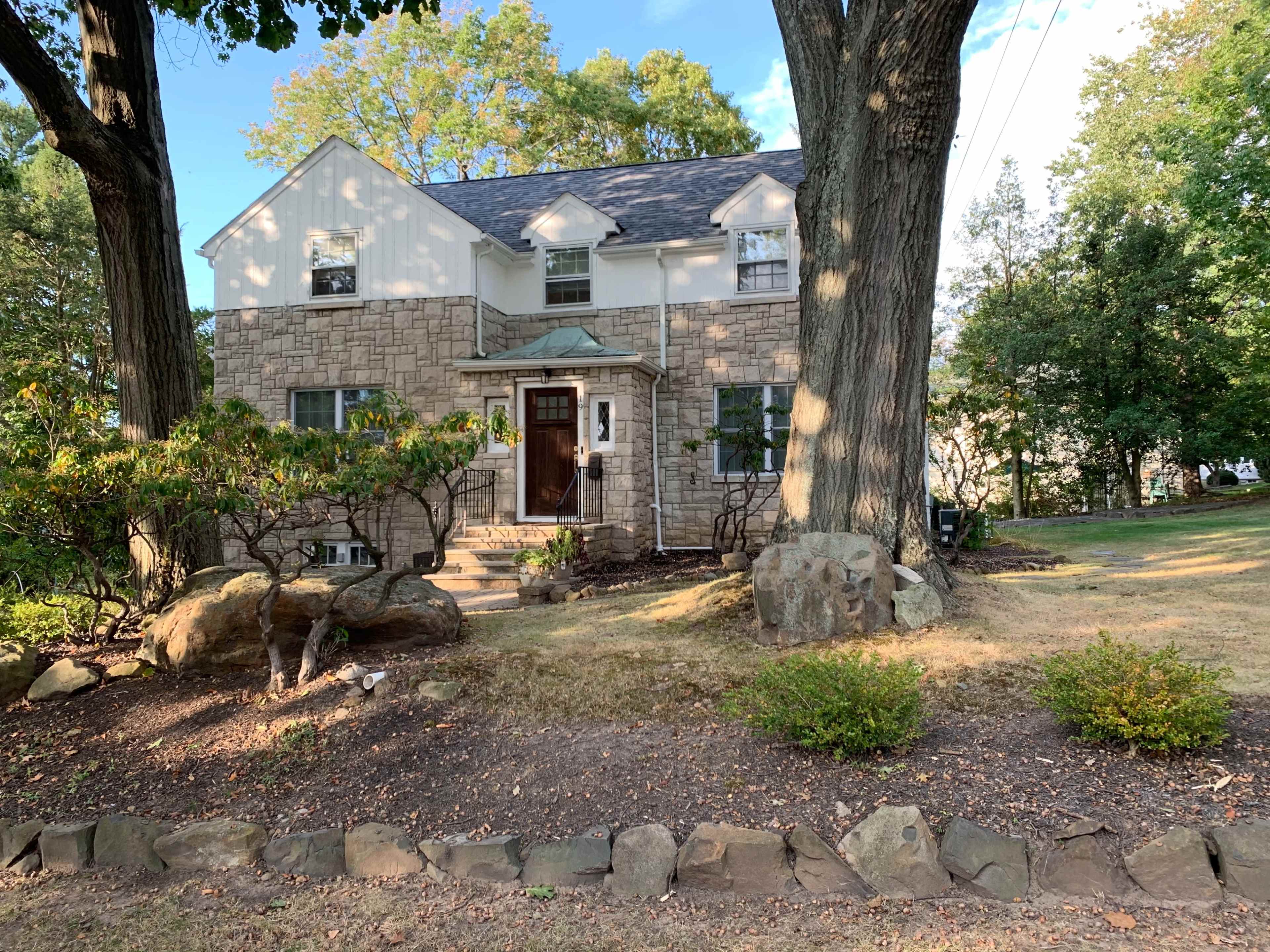 A two-story stone house with a sloped roof is surrounded by trees and landscaped bushes on a grassy yard.