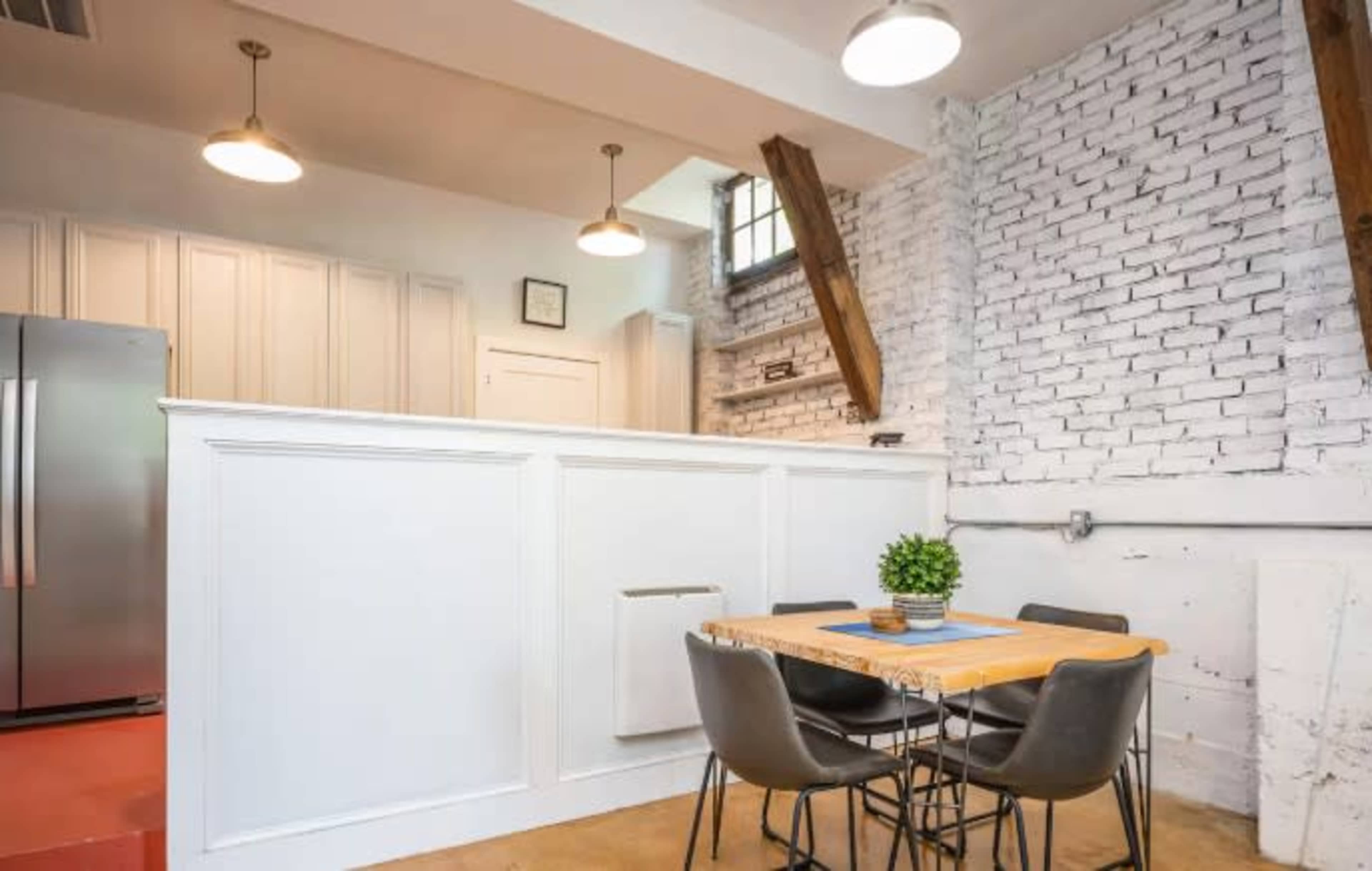 The image shows a kitchen area with a wooden dining table and chairs, a refrigerator, and exposed brick walls with wooden beams above.
