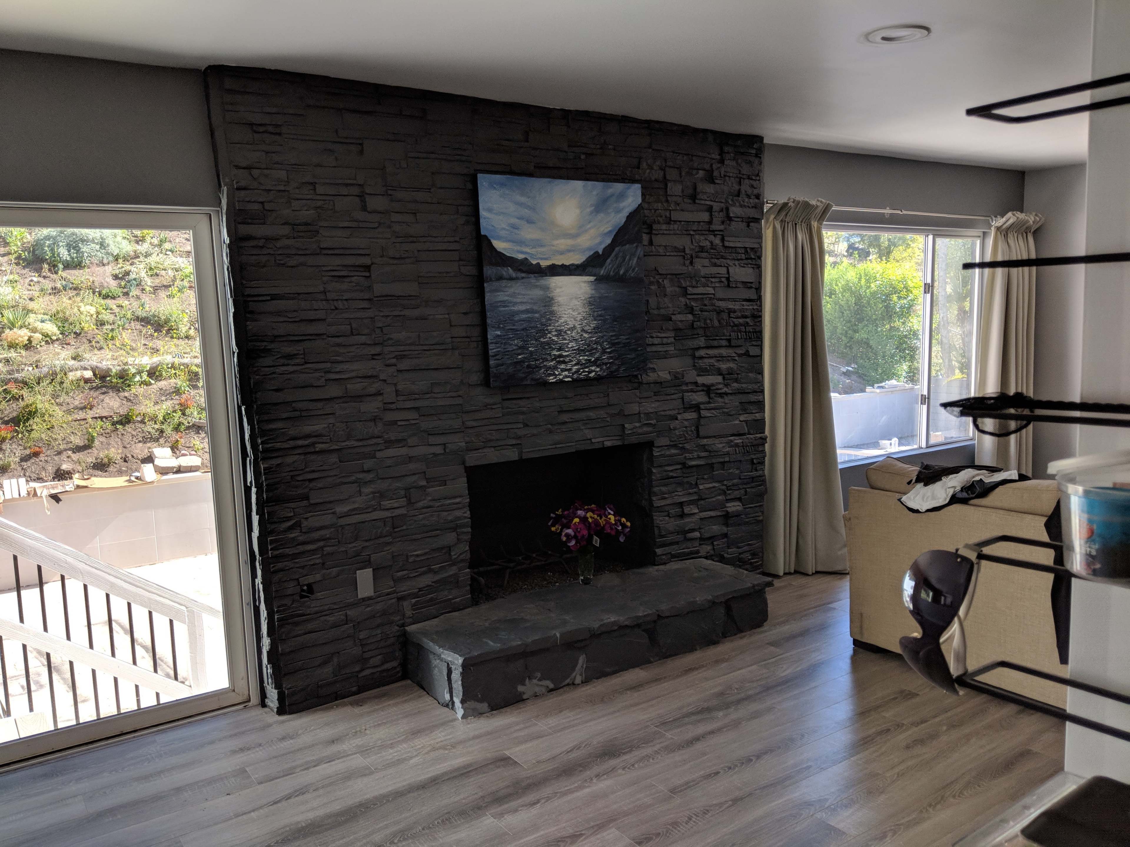 A living room with a stone fireplace, a mountain landscape painting above it, and large windows allowing natural light in.