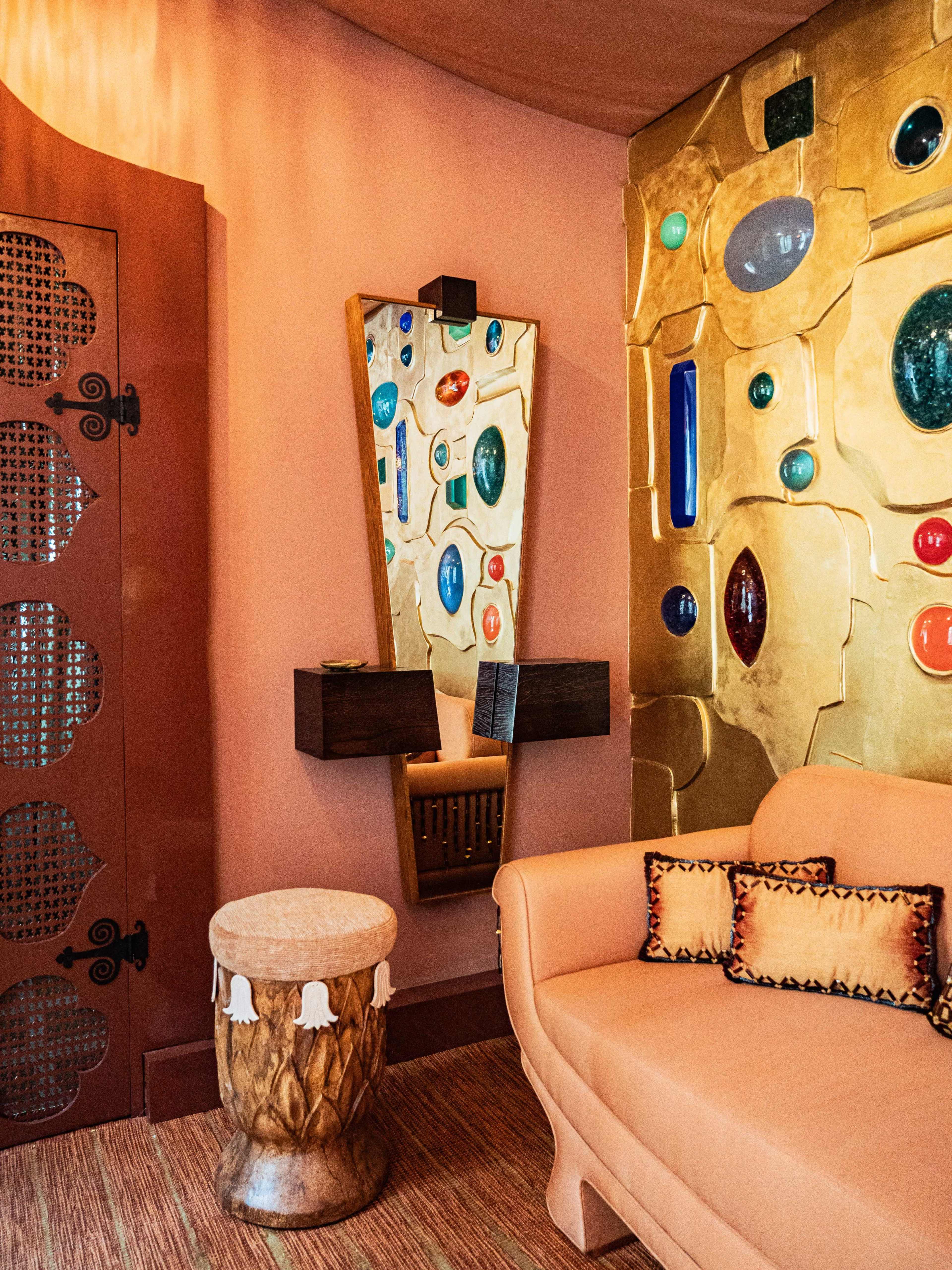 The image shows a room with a peach-colored wall adorned with colorful decorative stones, a wooden mirror, a small drum-shaped stool, and a beige sofa with patterned cushions.