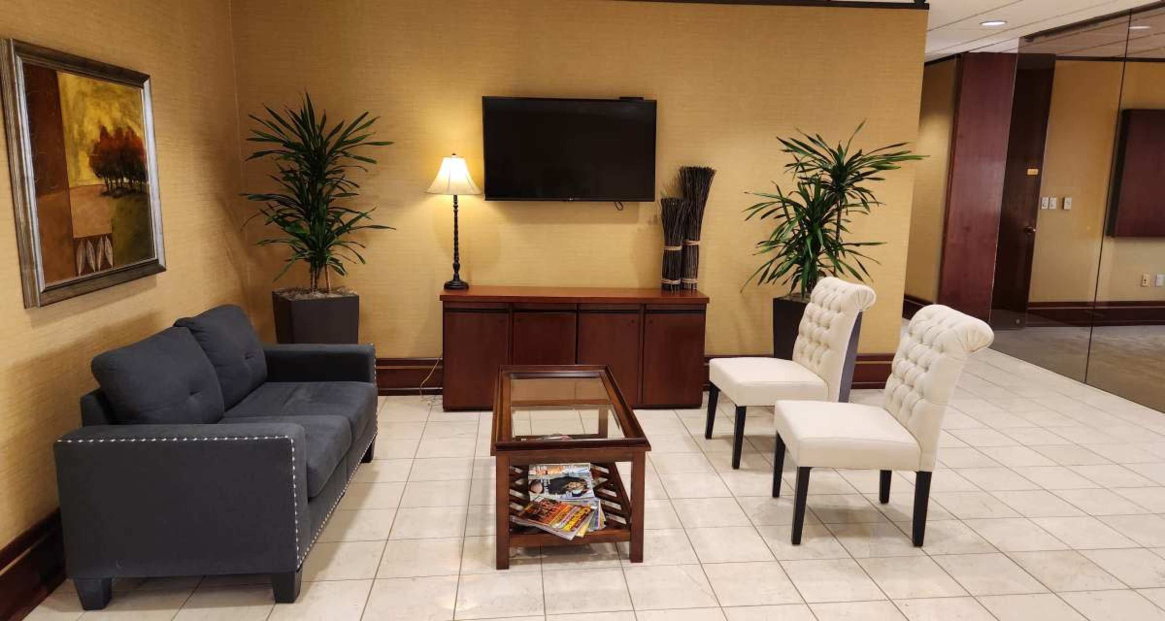 A waiting area with a gray sofa, two white chairs, a coffee table, a lamp, and indoor plants against a golden wall.