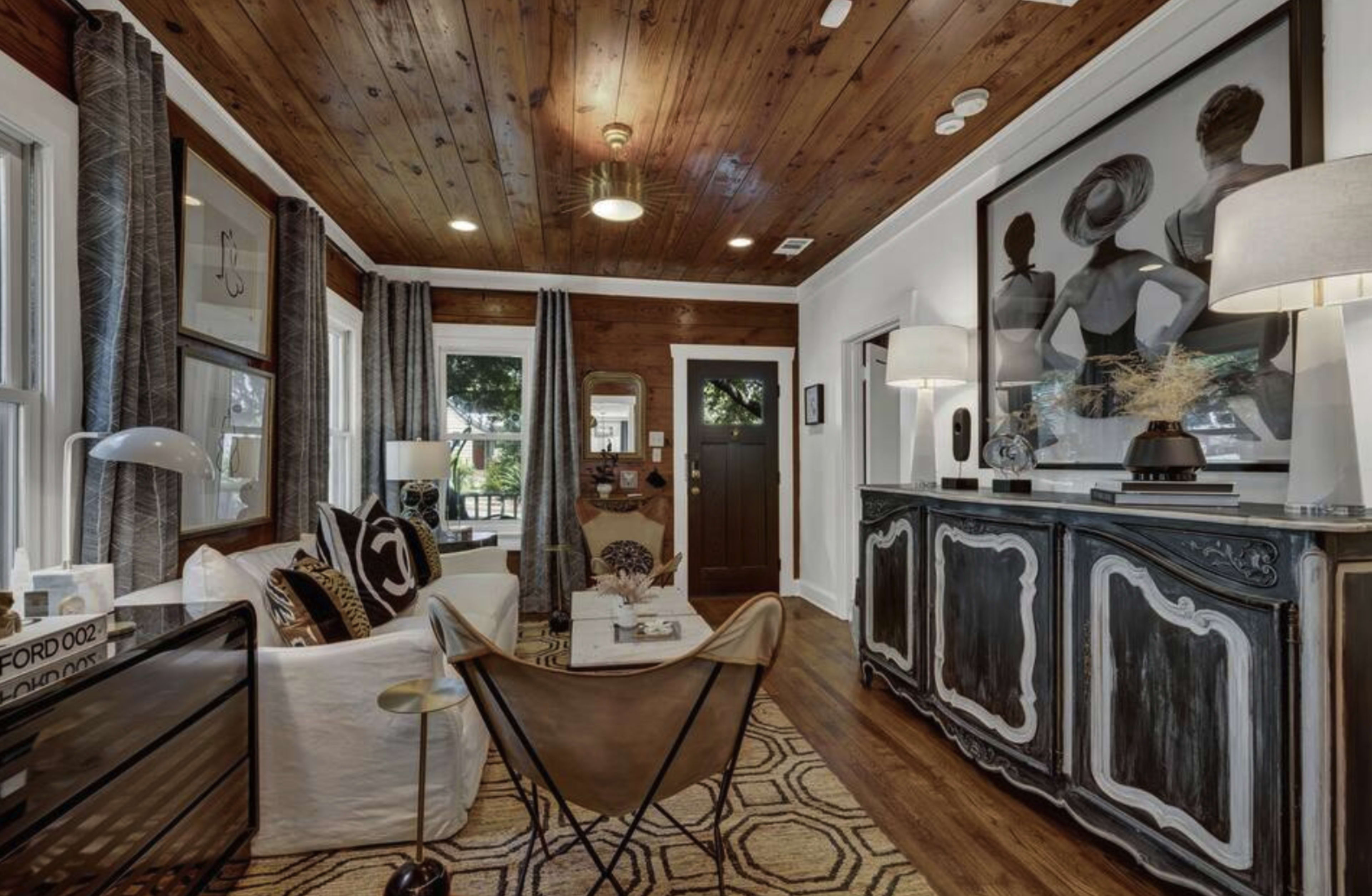 The image shows a cozy living room with wooden walls and ceiling, featuring a mix of modern and vintage furniture, including a leather butterfly chair and a decorative console.