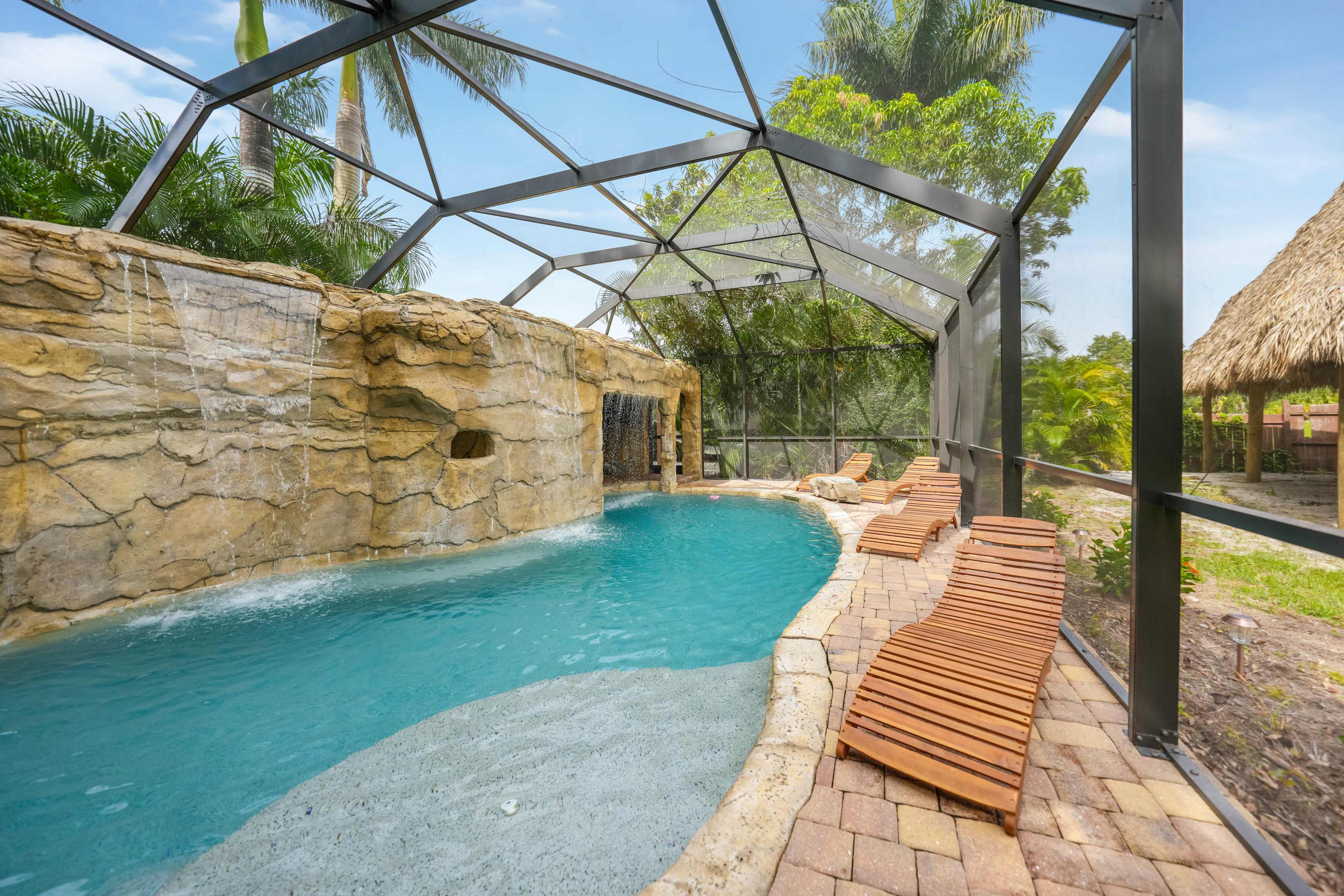 A pool area enclosed by a clear mesh screen, featuring a rock waterfall and wooden lounge chairs beside the water.