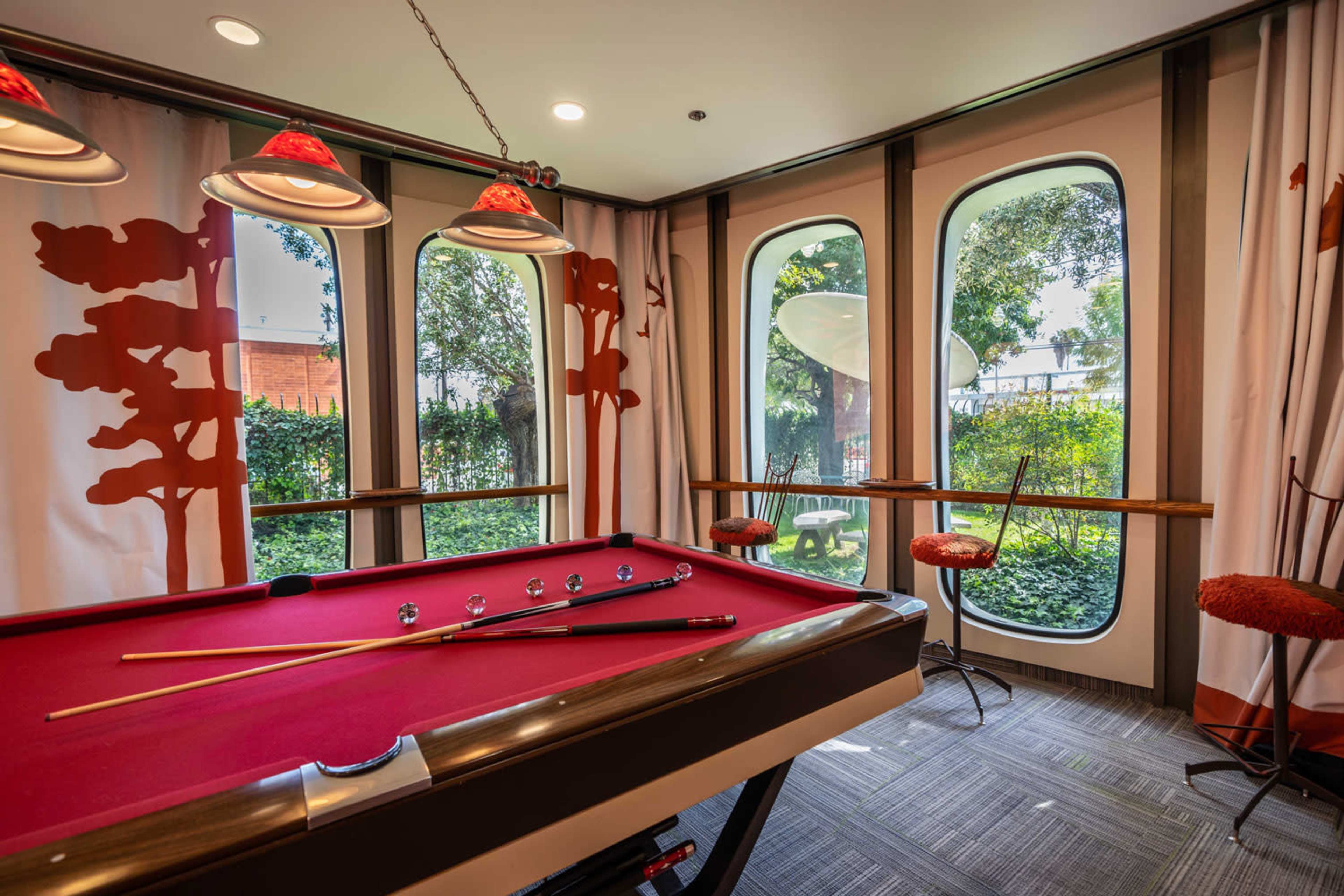 A brightly lit game room features a red pool table, surrounded by tall windows with views of greenery and stylish seating.
