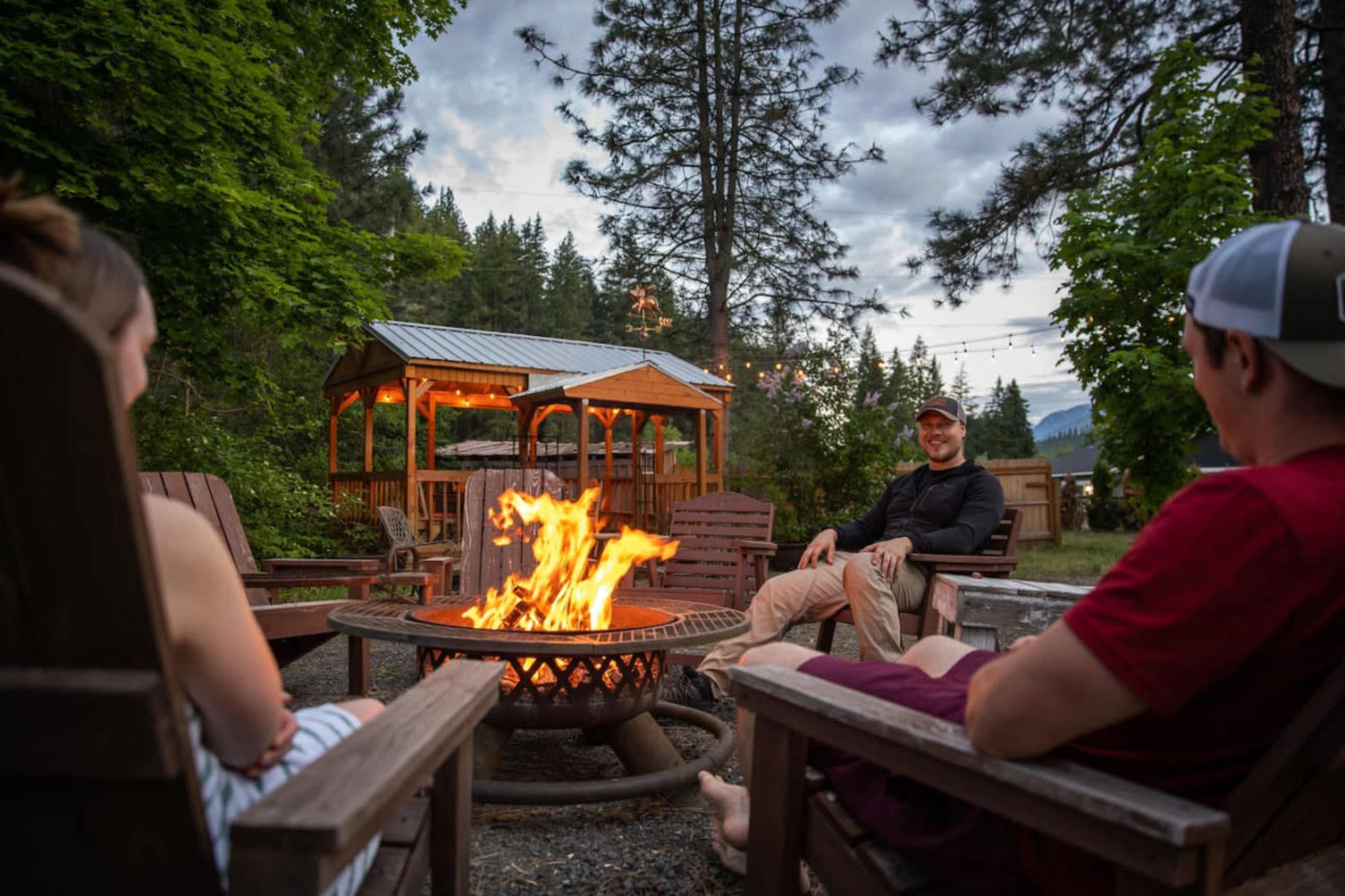 Three people sit around a fire pit, enjoying a campfire in an outdoor setting with trees and a gazebo in the background.