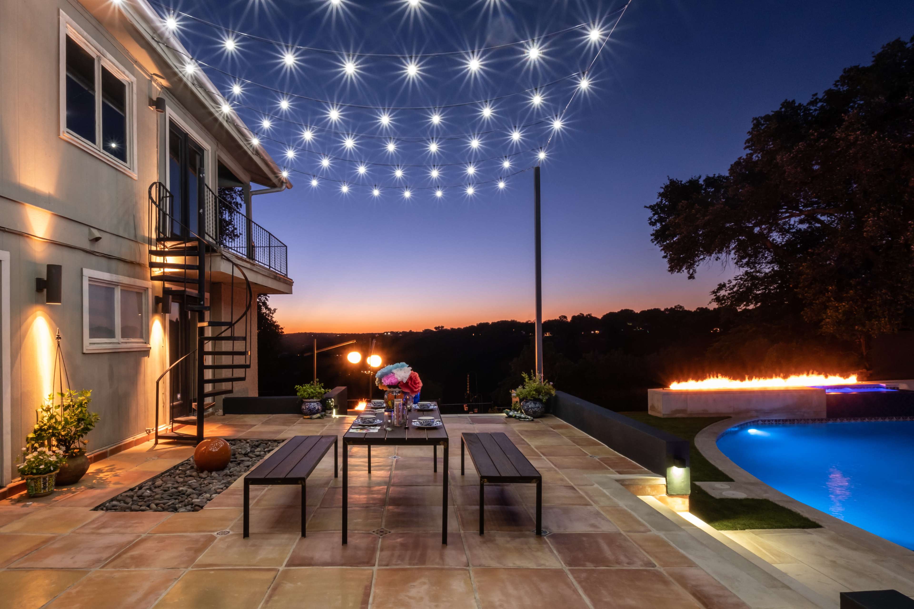 A patio with a dining setup is illuminated by string lights at dusk, featuring a pool and a fire feature in the background.