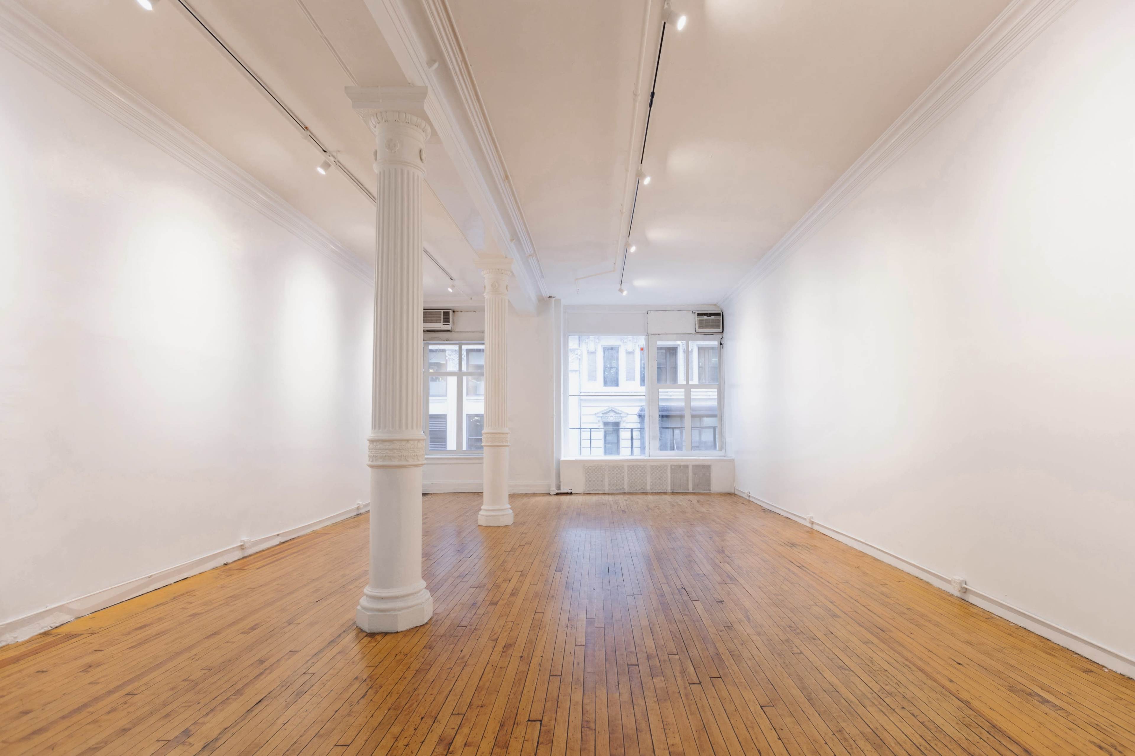 The image shows a spacious, empty room with wooden flooring, white walls, and large windows allowing natural light.
