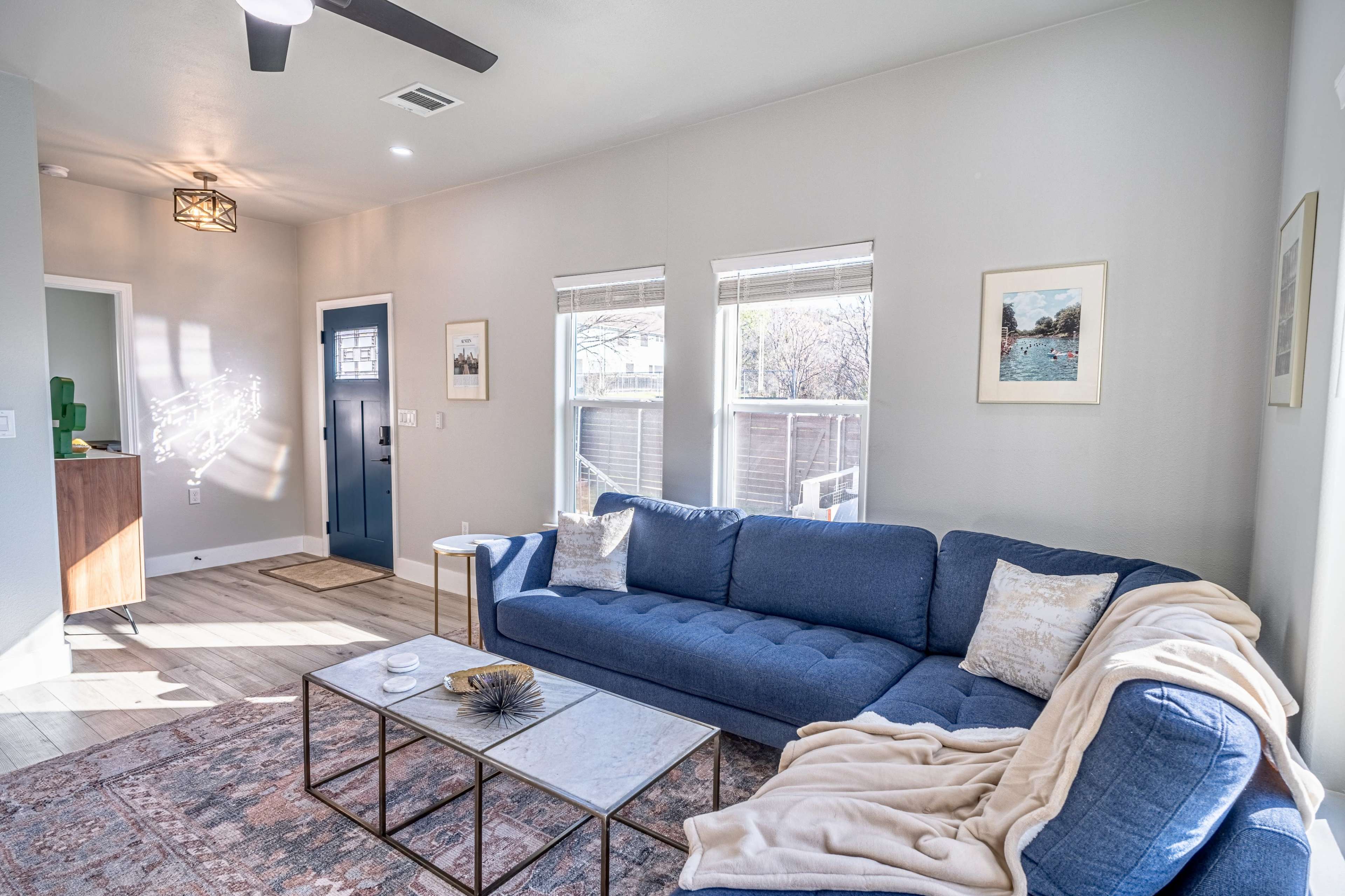 The image shows a living room with a blue sofa, a coffee table, and two windows allowing natural light to fill the space.