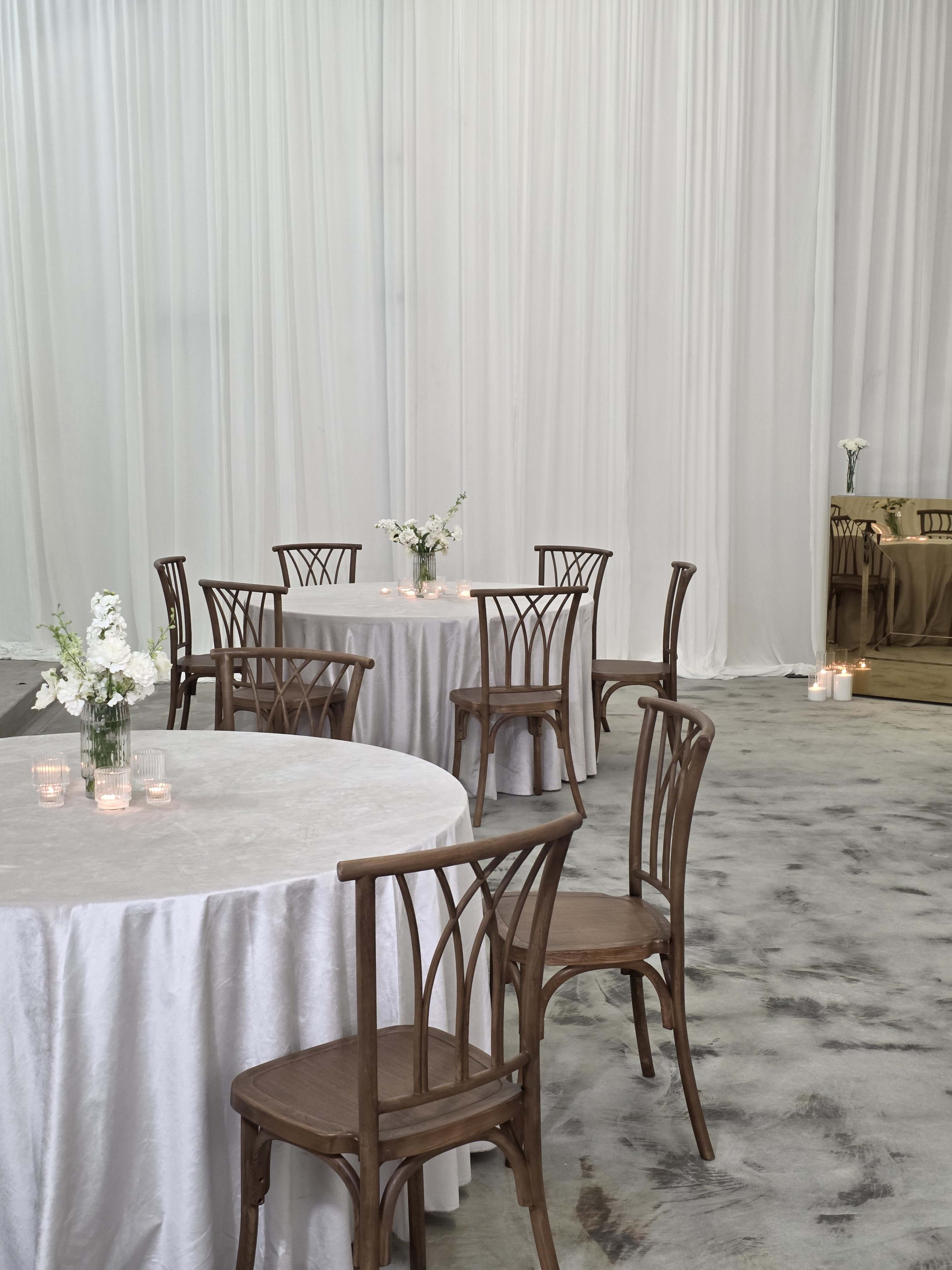 A spacious venue features round tables covered with light tablecloths, surrounded by wooden chairs, and adorned with flower arrangements and candles.
