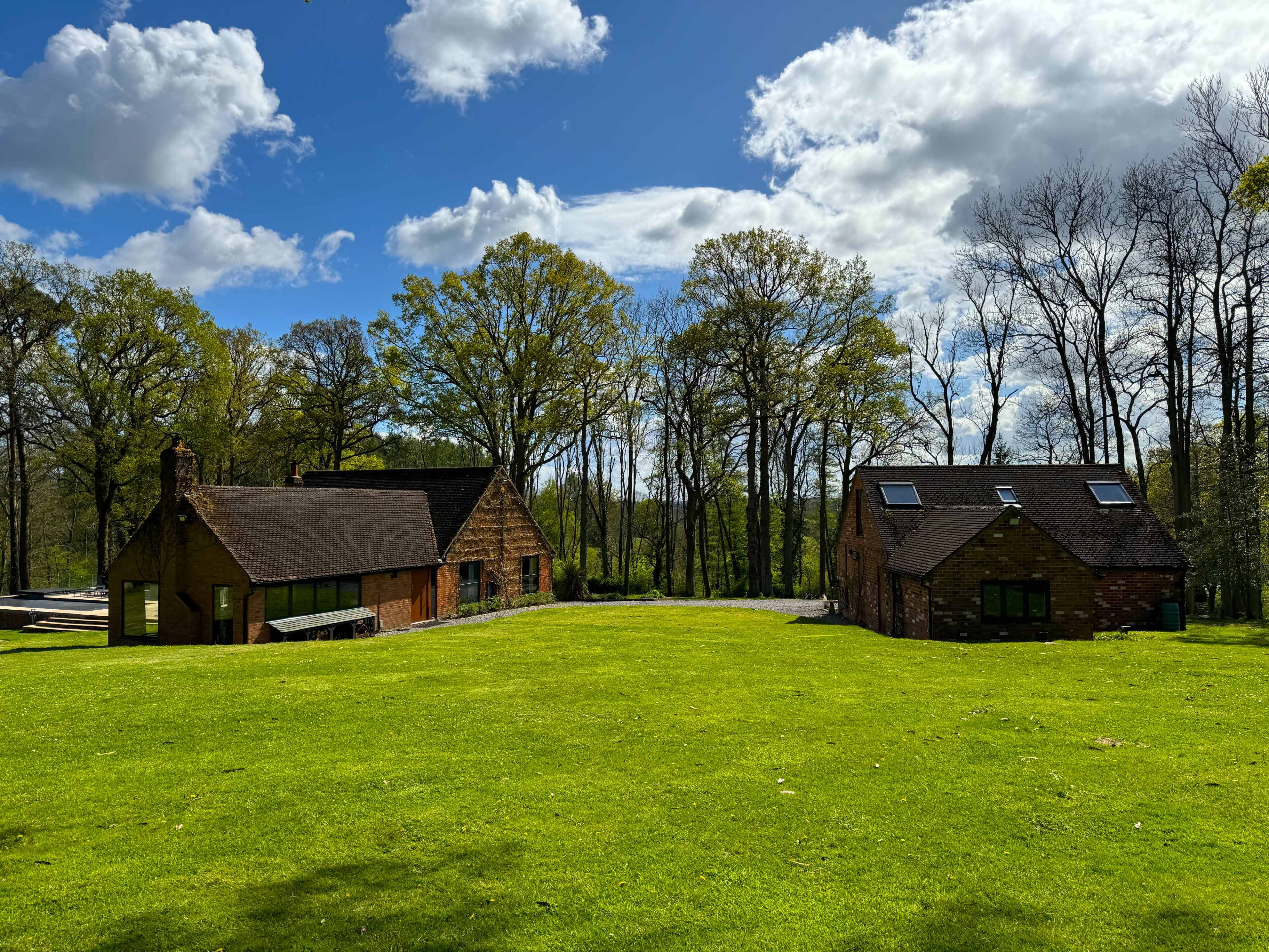 Two rustic houses with sloped roofs sit on a green lawn surrounded by tall trees and a blue sky with white clouds.