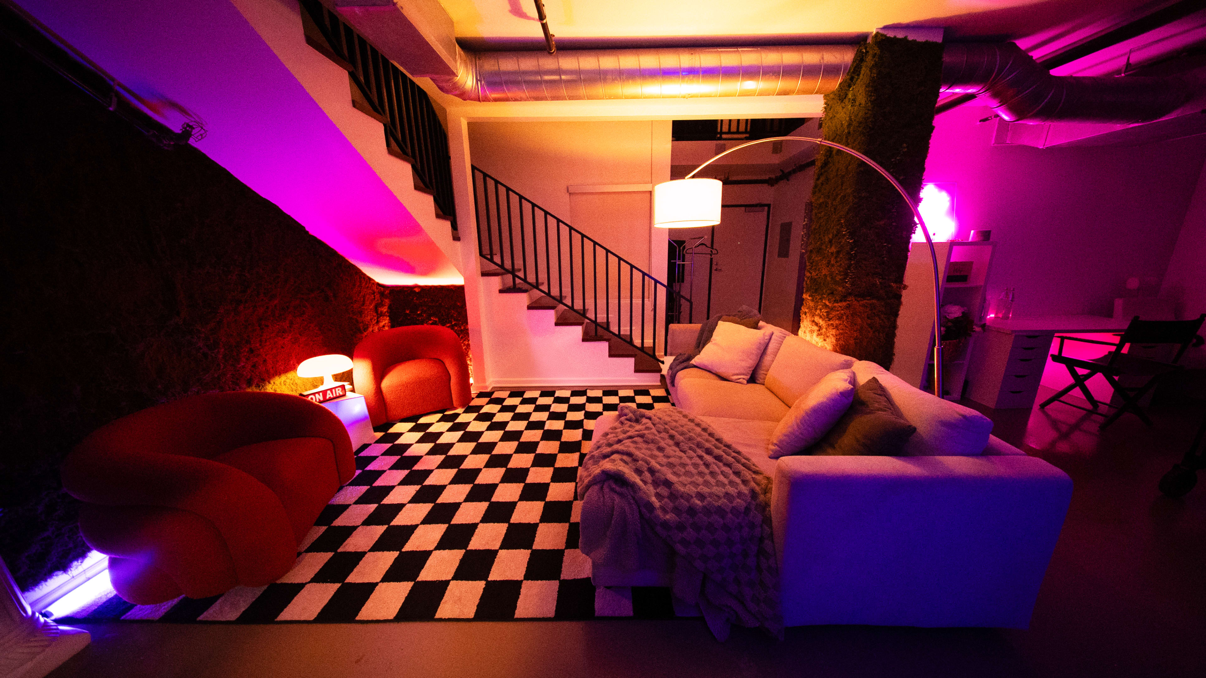 The image shows a modern interior space featuring a checkered rug, a white sofa, and two red armchairs, illuminated by purple and pink lighting.