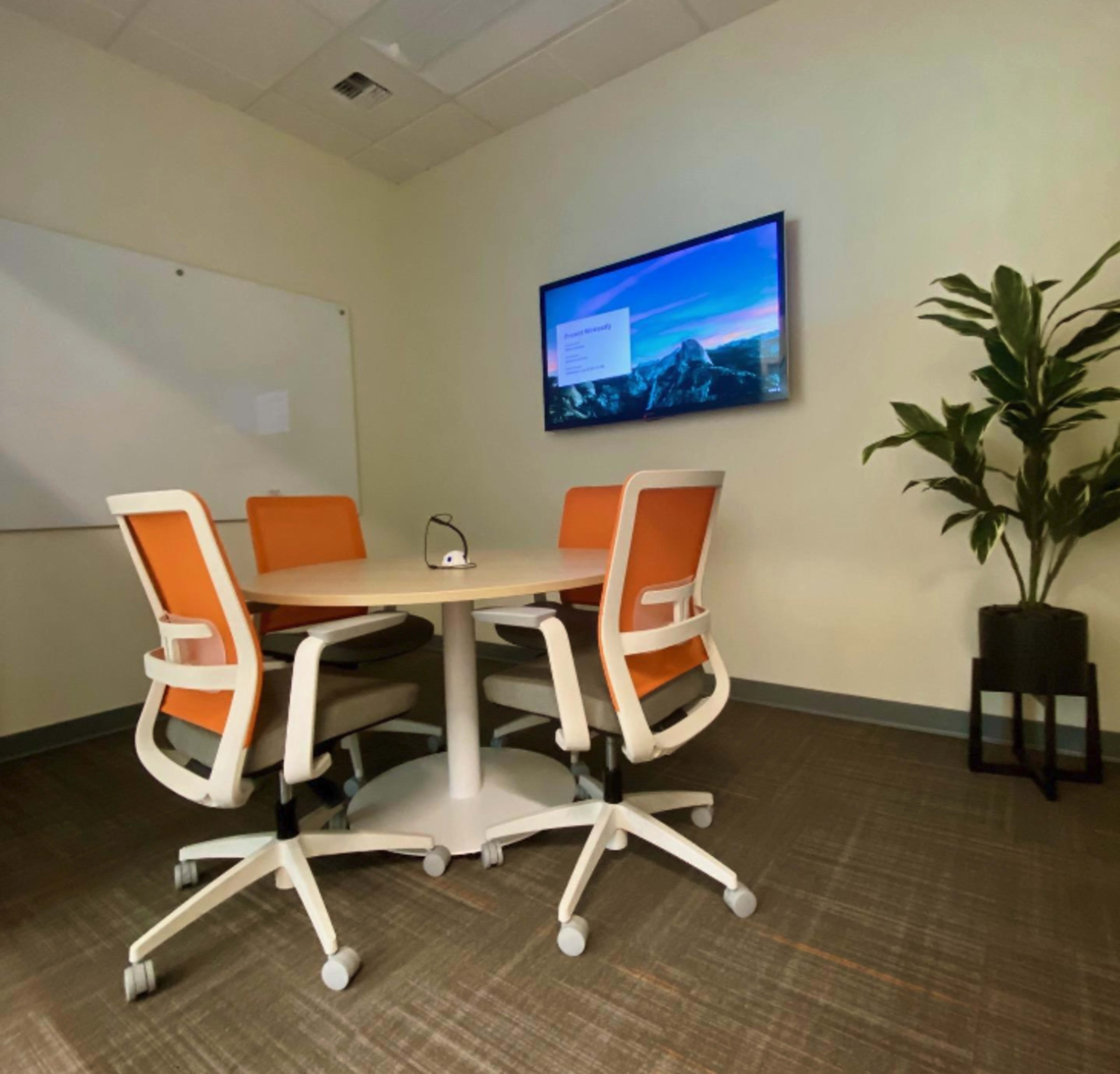 A small conference room features a round table surrounded by four chairs with orange cushions, a wall-mounted screen displaying a presentation, and a potted plant in the corner.
