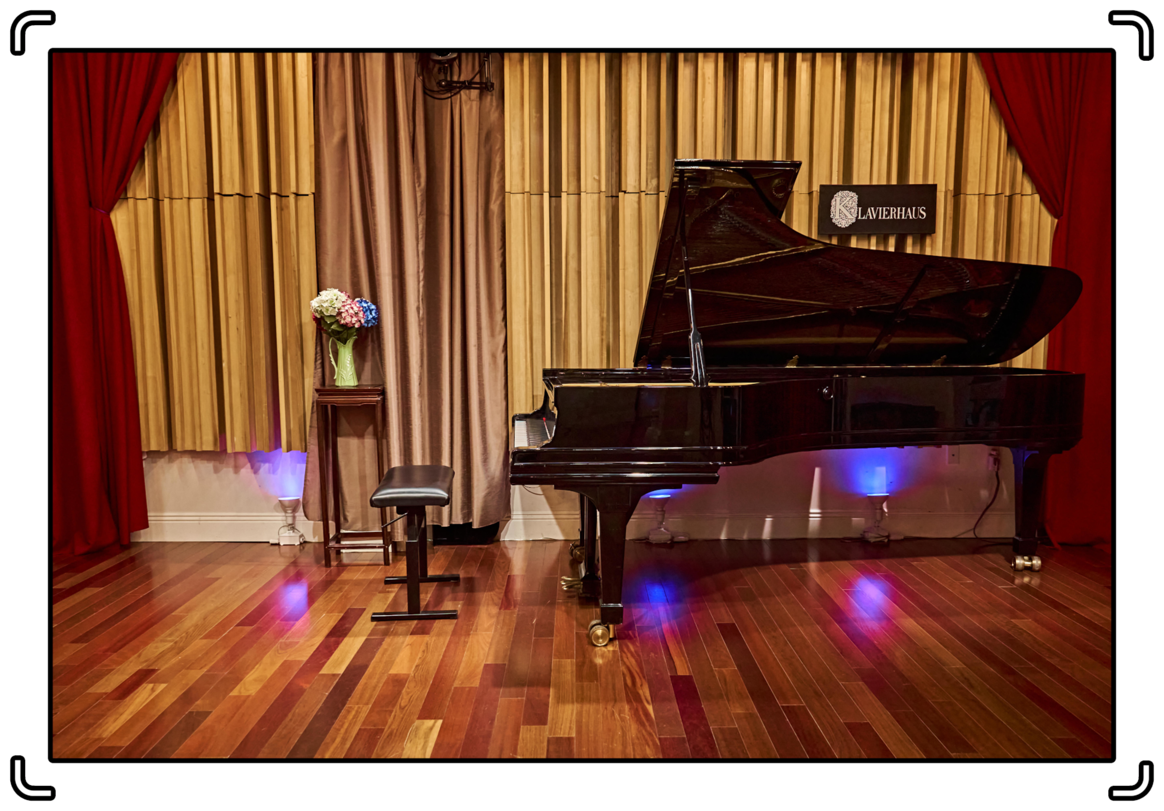 A black grand piano stands beside a stool on a polished wooden floor, with a vase of flowers on a nearby table and soundproof curtains in the background.