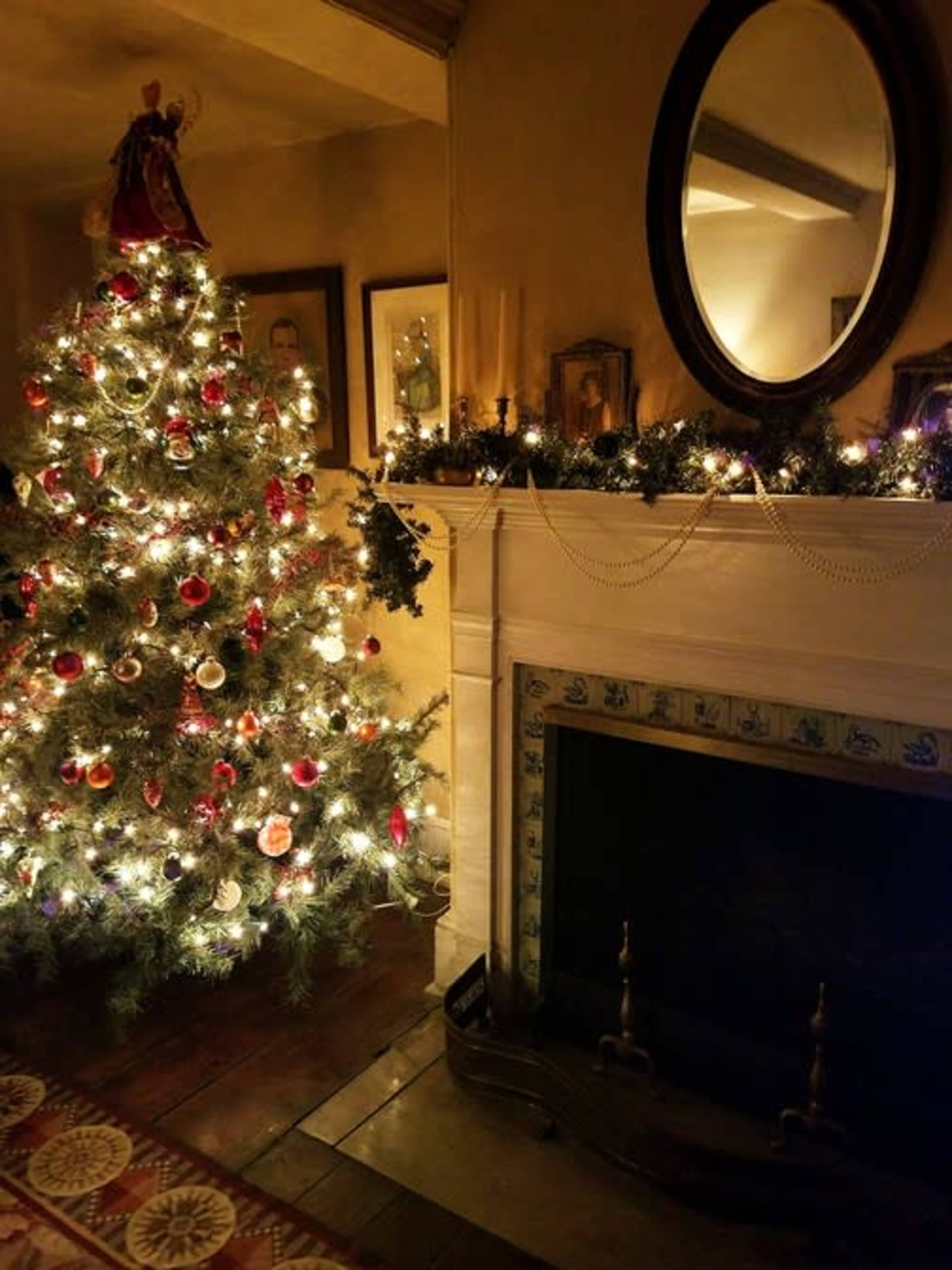 A decorated Christmas tree with lights and ornaments stands next to a fireplace adorned with garlands and framed pictures on the wall.