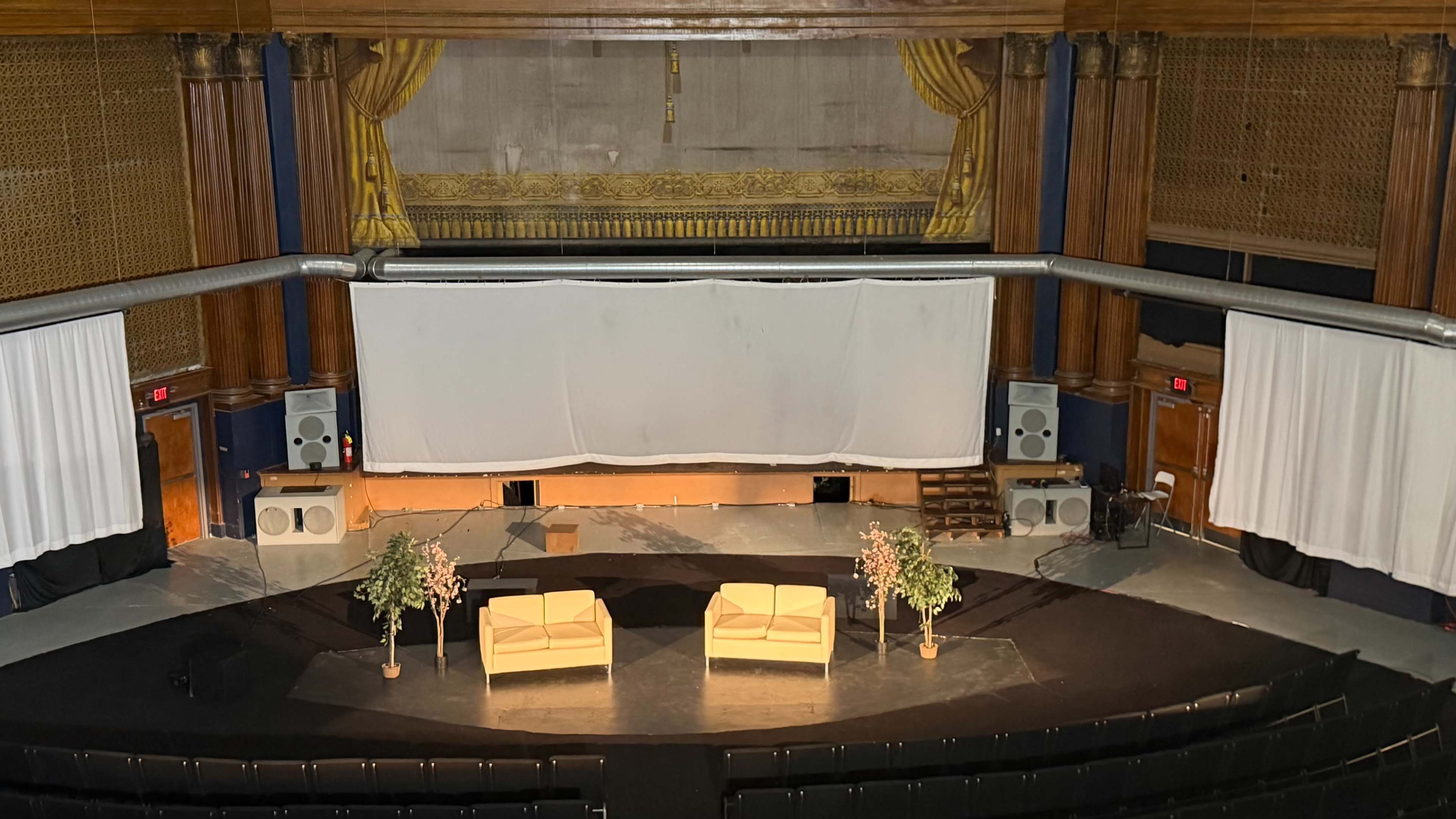 The image shows a theater stage featuring two light-colored chairs, two plants, and a large white screen at the back.