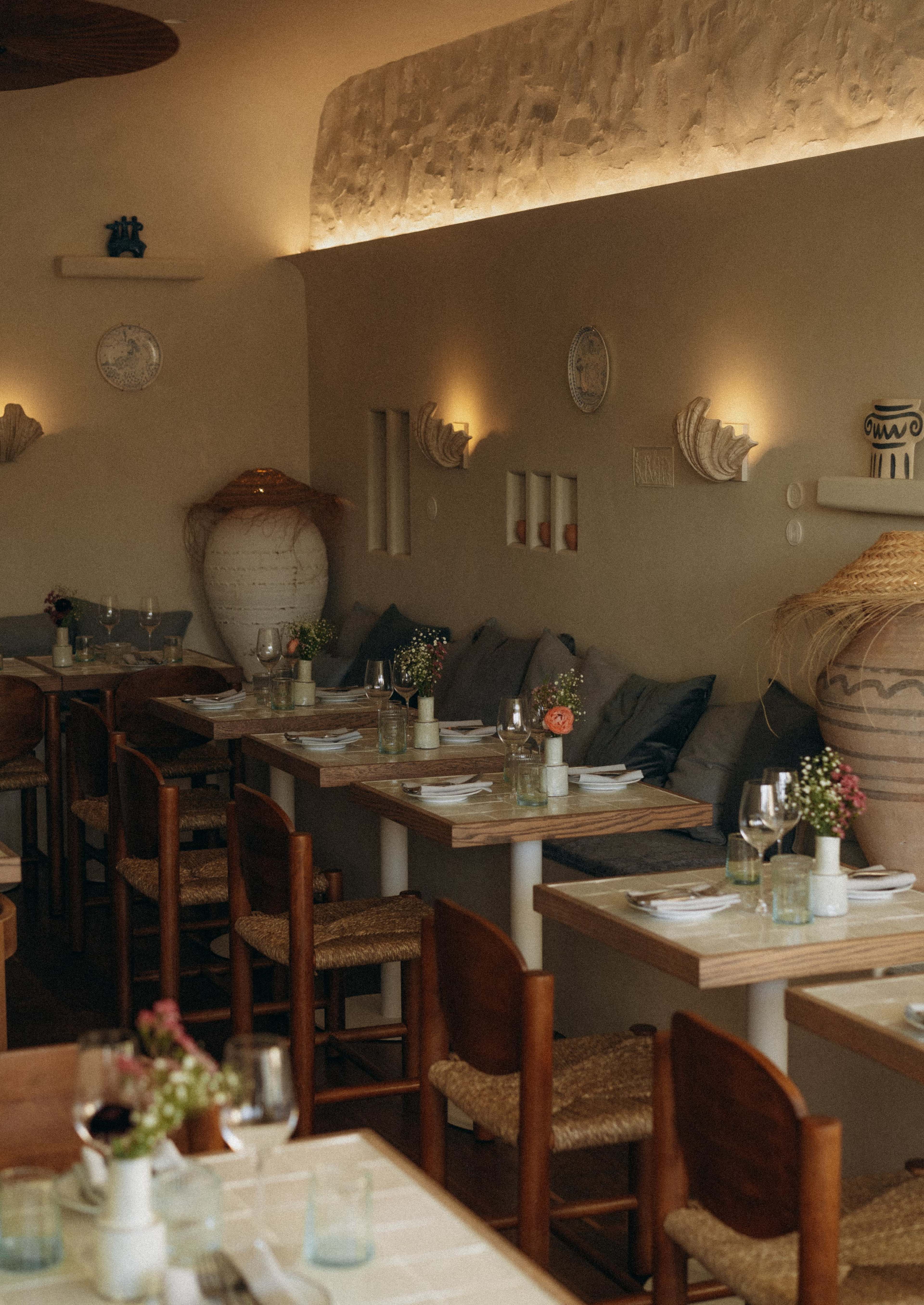 The image shows a cozy restaurant interior with wooden tables, woven chairs, and minimalist decor featuring plants and pottery.