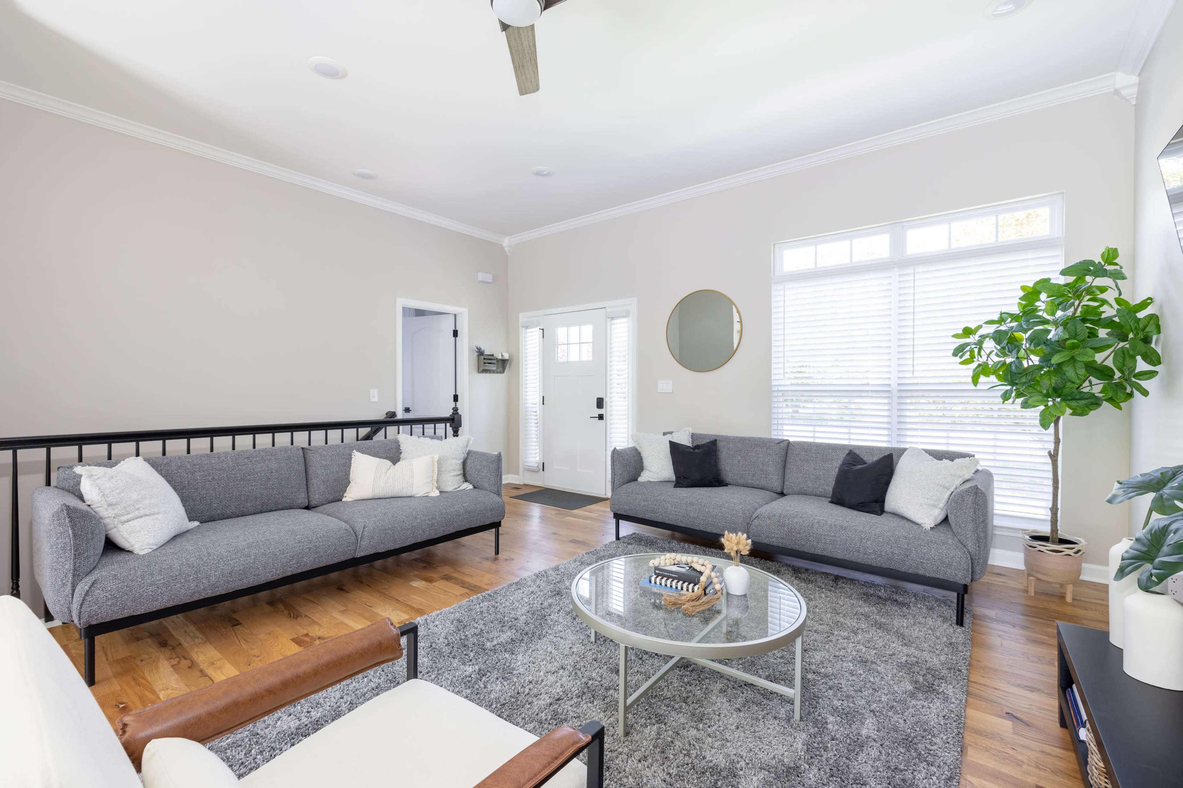 The living room features two gray sofas, a round glass coffee table, a rug, and a large window with white blinds.
