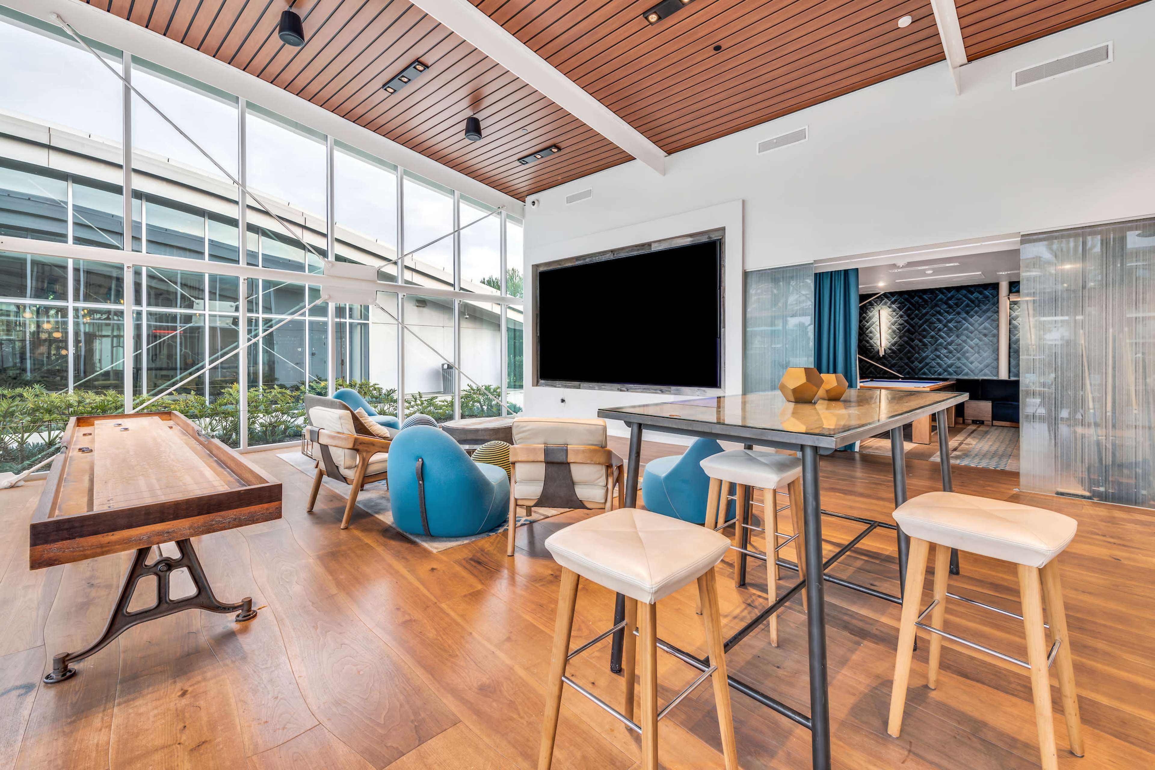 The image shows a modern lounge area featuring a large screen, a wooden table, several stools, and bean bag chairs, with large windows allowing natural light.