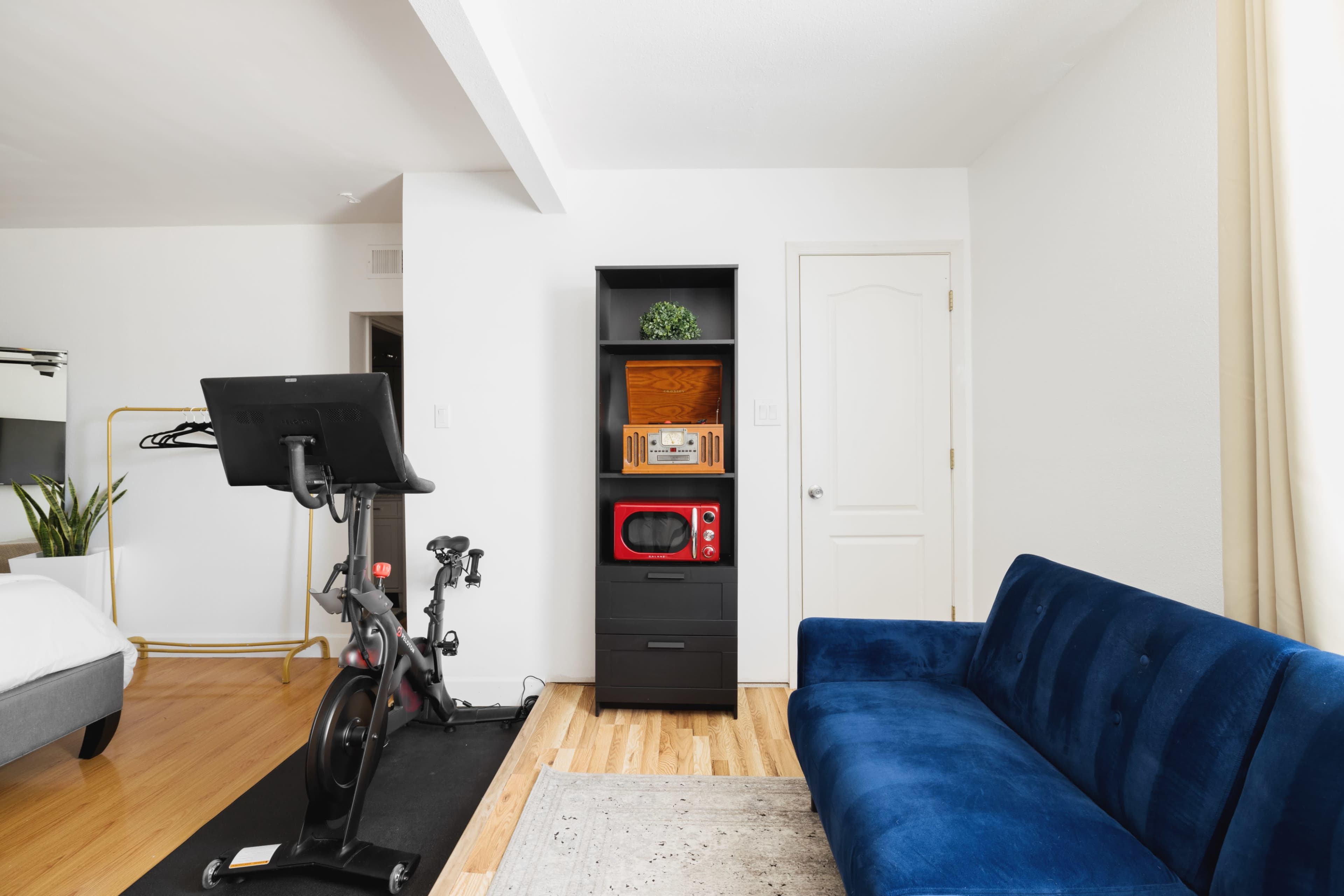 The image shows a small living space featuring a blue couch, a black shelving unit with a microwave, a stationary exercise bike, and a door leading to another room.