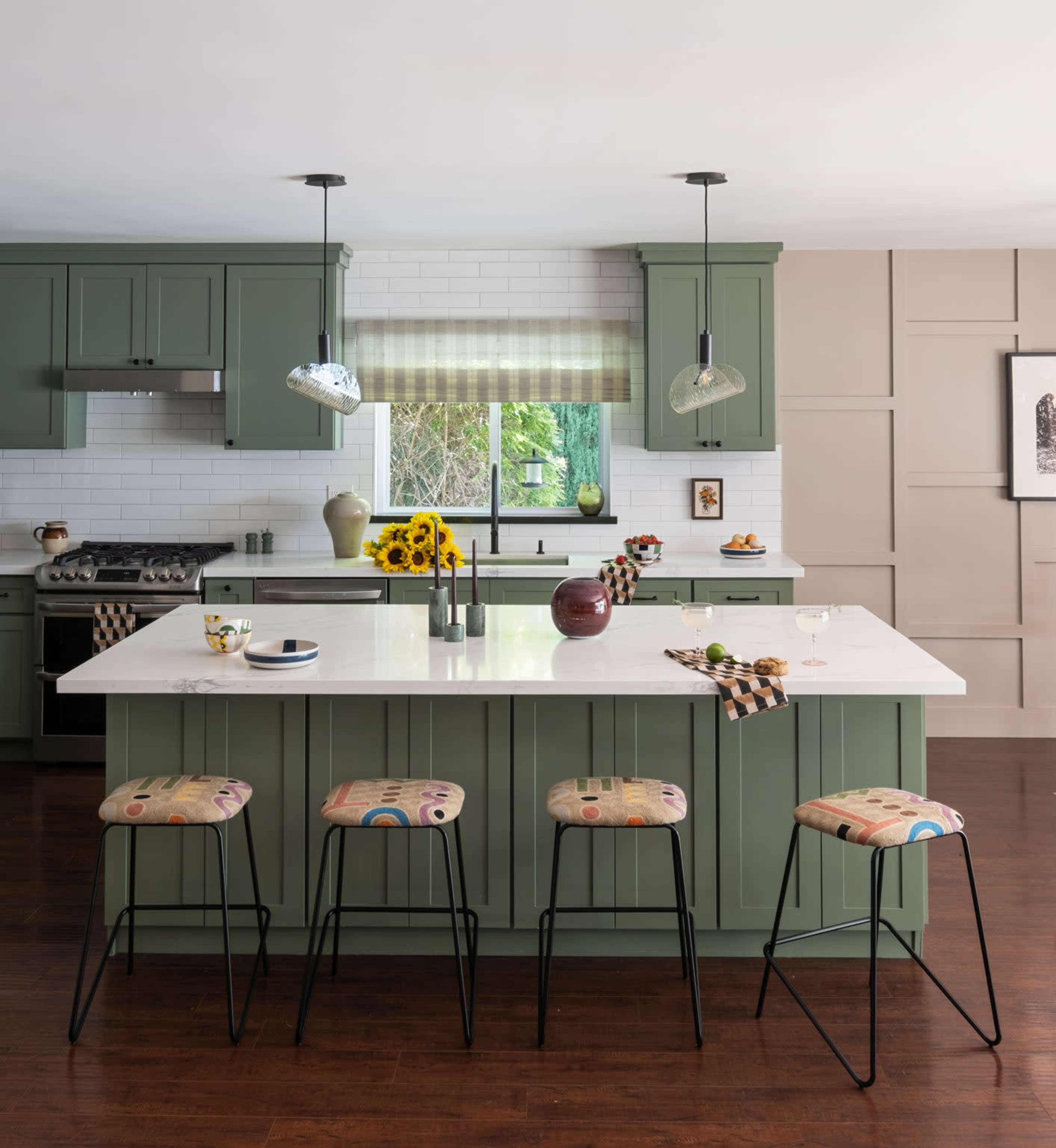 The kitchen features green cabinetry, a large white island with bar stools, and a window overlooking a garden filled with sunflowers.