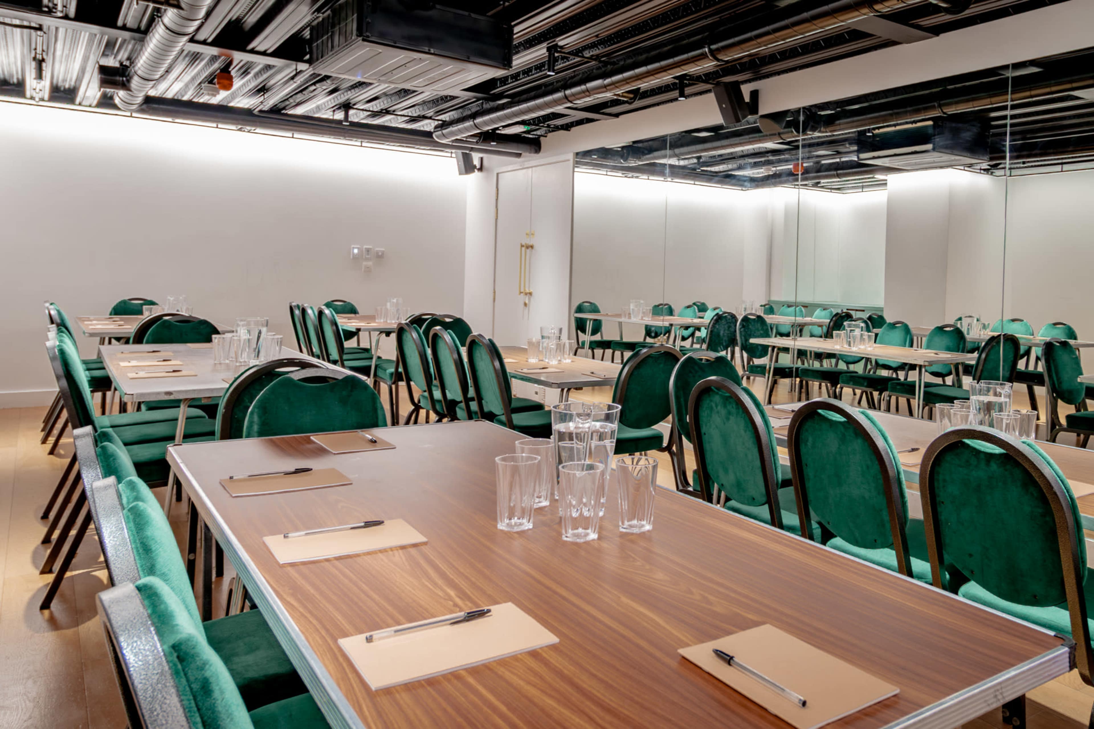 The image shows a modern conference room with multiple wooden tables set for a meeting, featuring green upholstered chairs, notepads, and glasses of water.