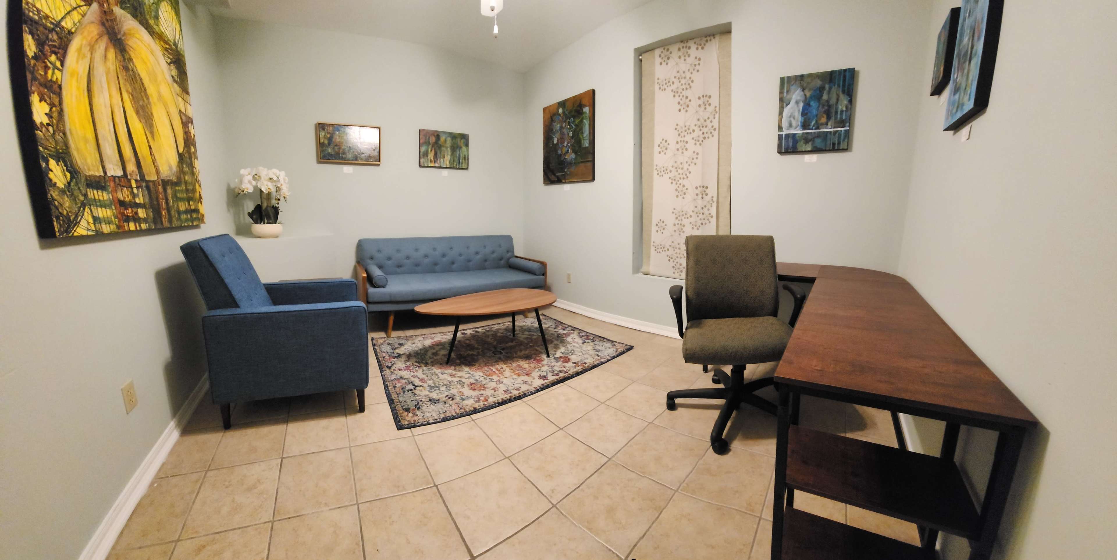 A small, well-lit waiting room with two blue couches, a round coffee table, a patterned rug, a desk with a chair, and several framed paintings on the walls.