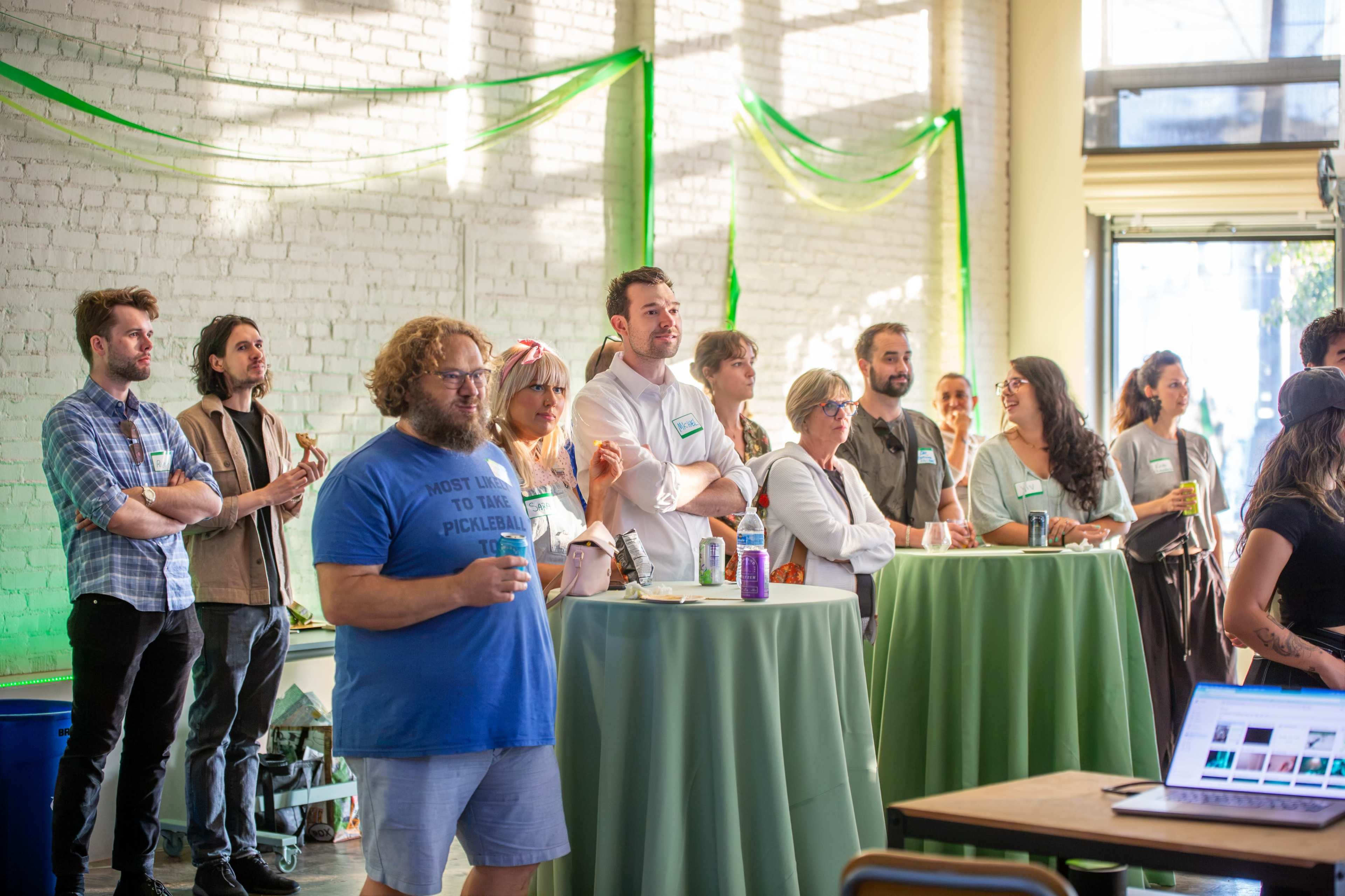 A group of people stands in a brightly lit room, engaged in conversation and mingling around tall tables adorned with green tablecloths.