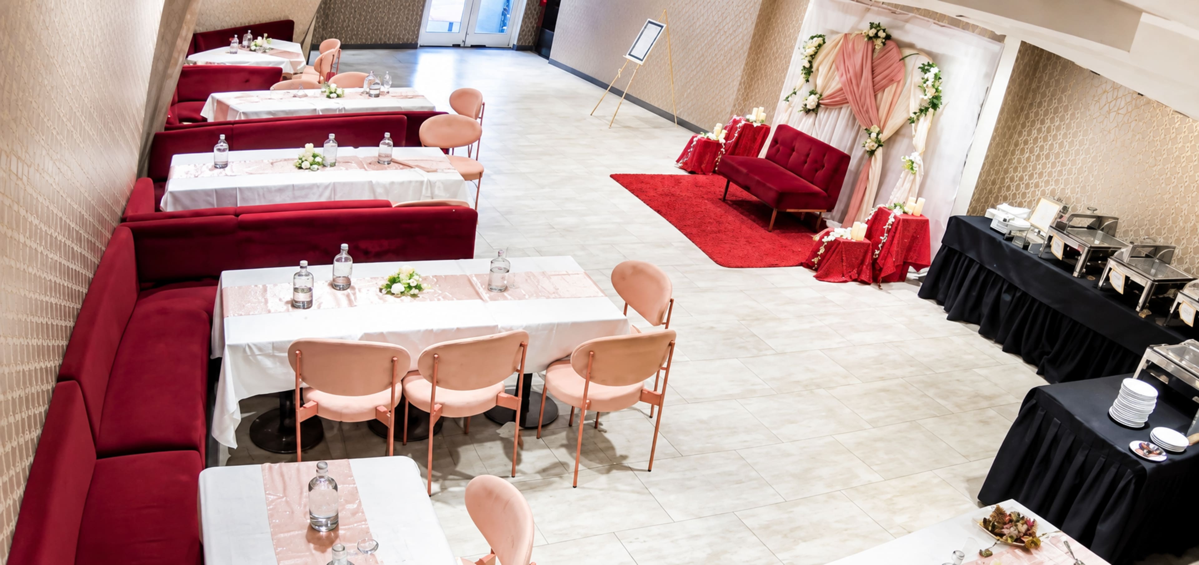 The image shows a banquet hall arranged with several tables covered in white cloth, pink chairs, and a decorative area featuring a backdrop of draped fabric and floral arrangements.